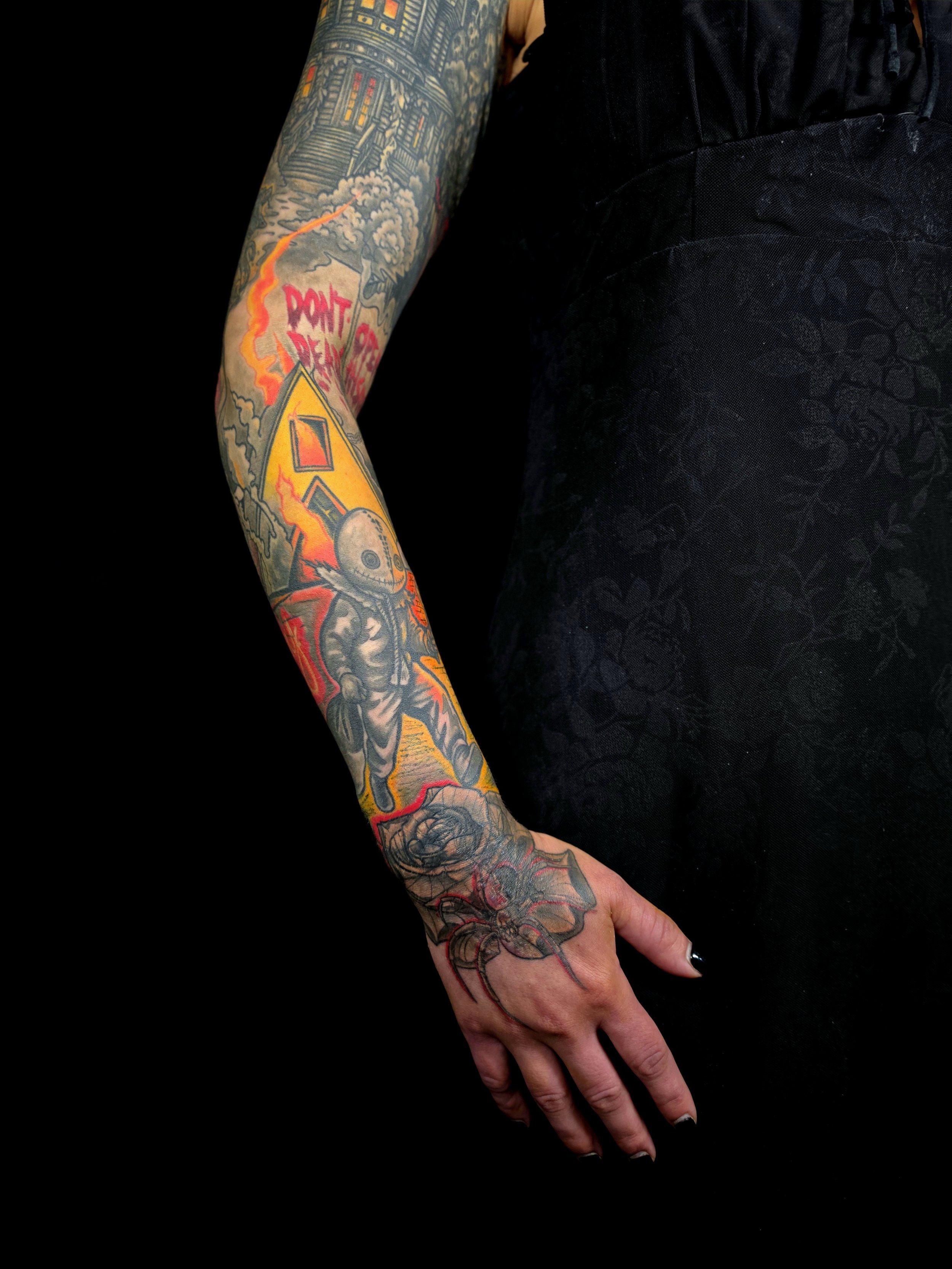 A person's arm with a colorful sleeve tattoo, including a skull and flames, resting on their lap against a black background.