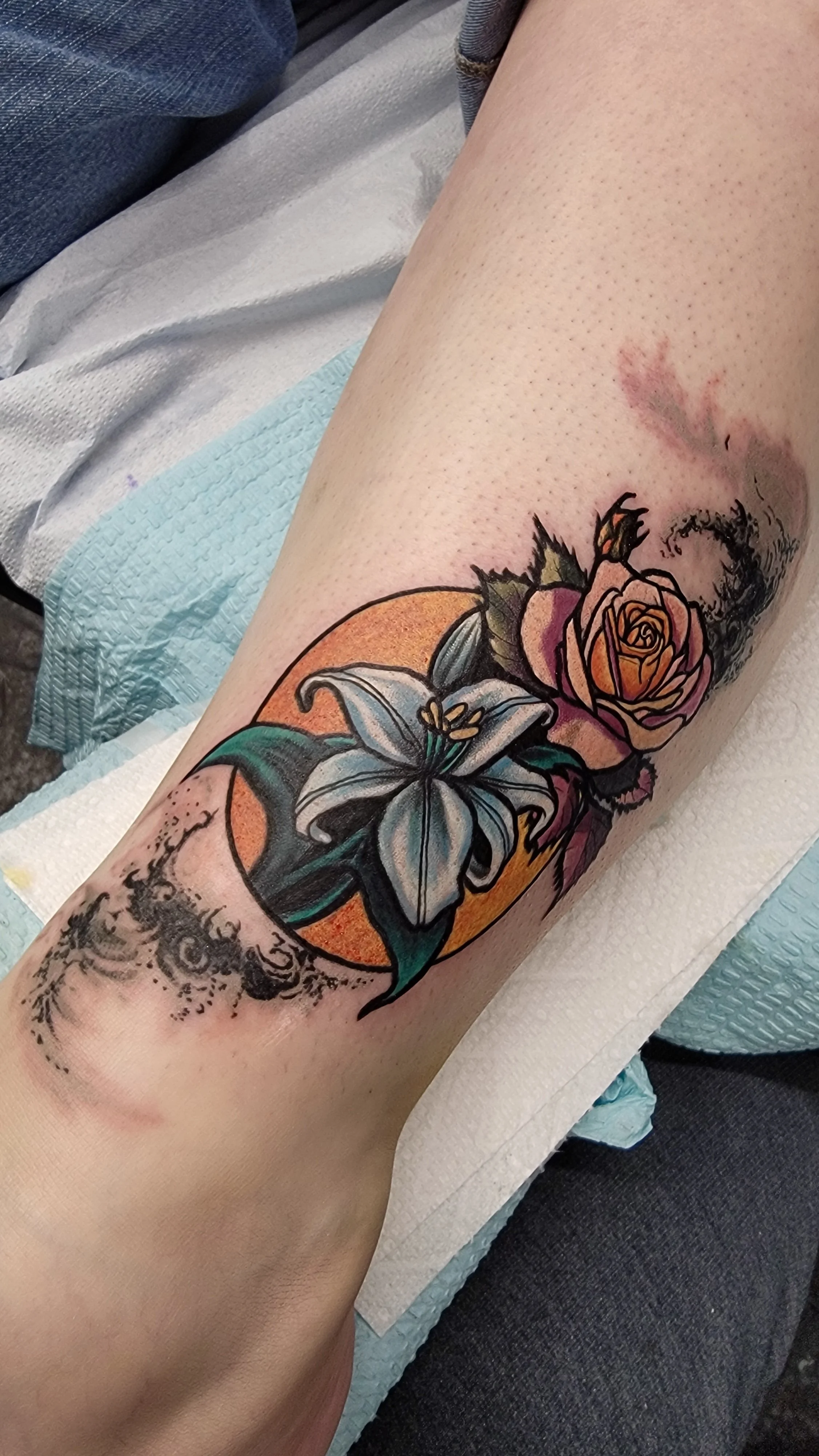 Colorful tattoo of a white lily and an orange rose on a person's forearm, with a background of an orange circle and a black and gray decorative pattern.