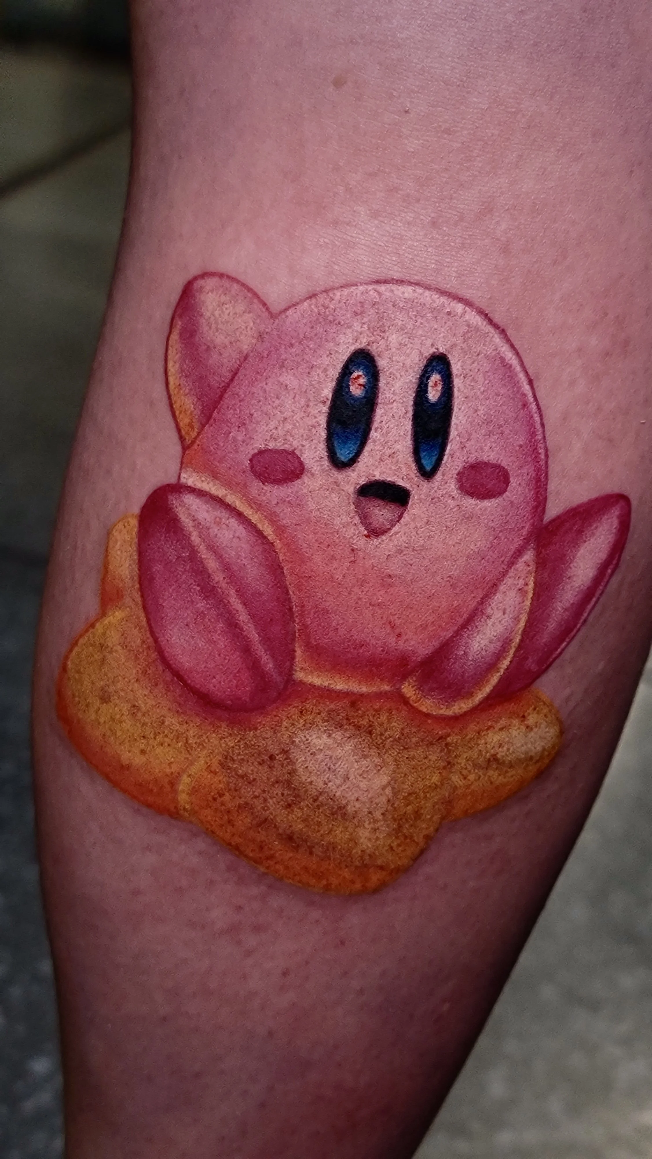 Colorful tattoo of Kirby, a pink video game character, sitting on an orange patch of ground.
