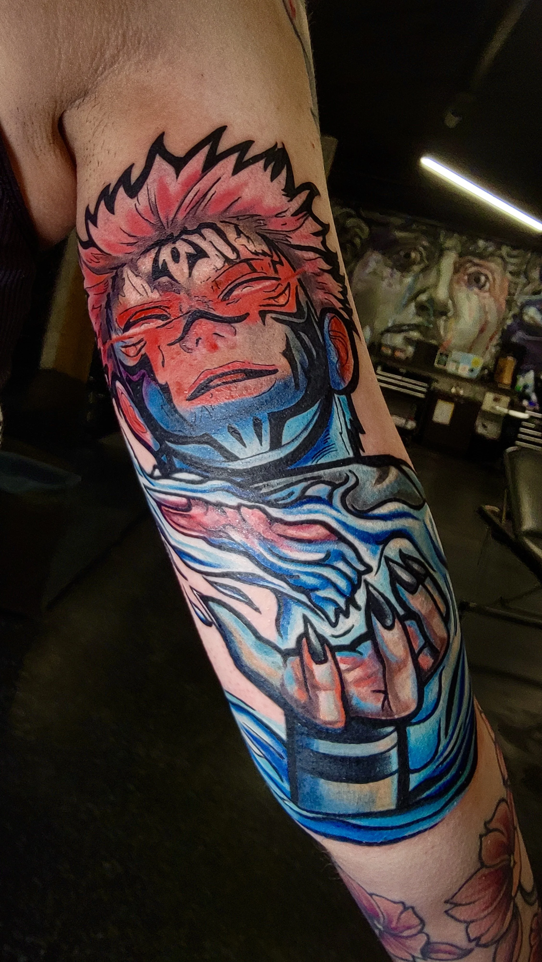 Colorful tattoo of a person's face with spiky hair and closed eyes on person's upper arm, with water and wave designs below, in an art studio with murals in the background.