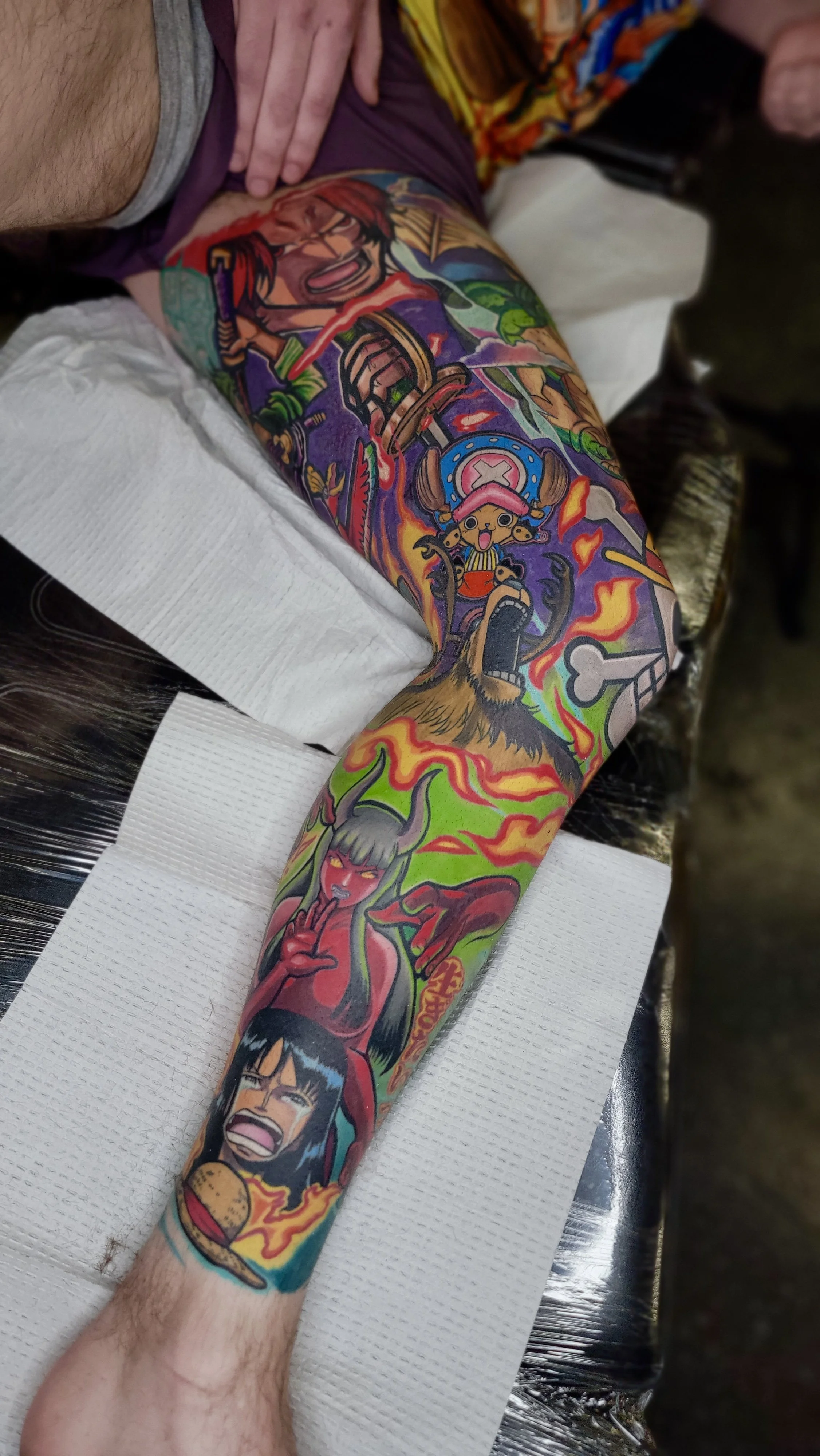 Colorful tattoo sleeve featuring cartoon characters and vibrant imagery, including a fierce warrior, a happy character with a hat, a demon girl, and various flames and symbols.