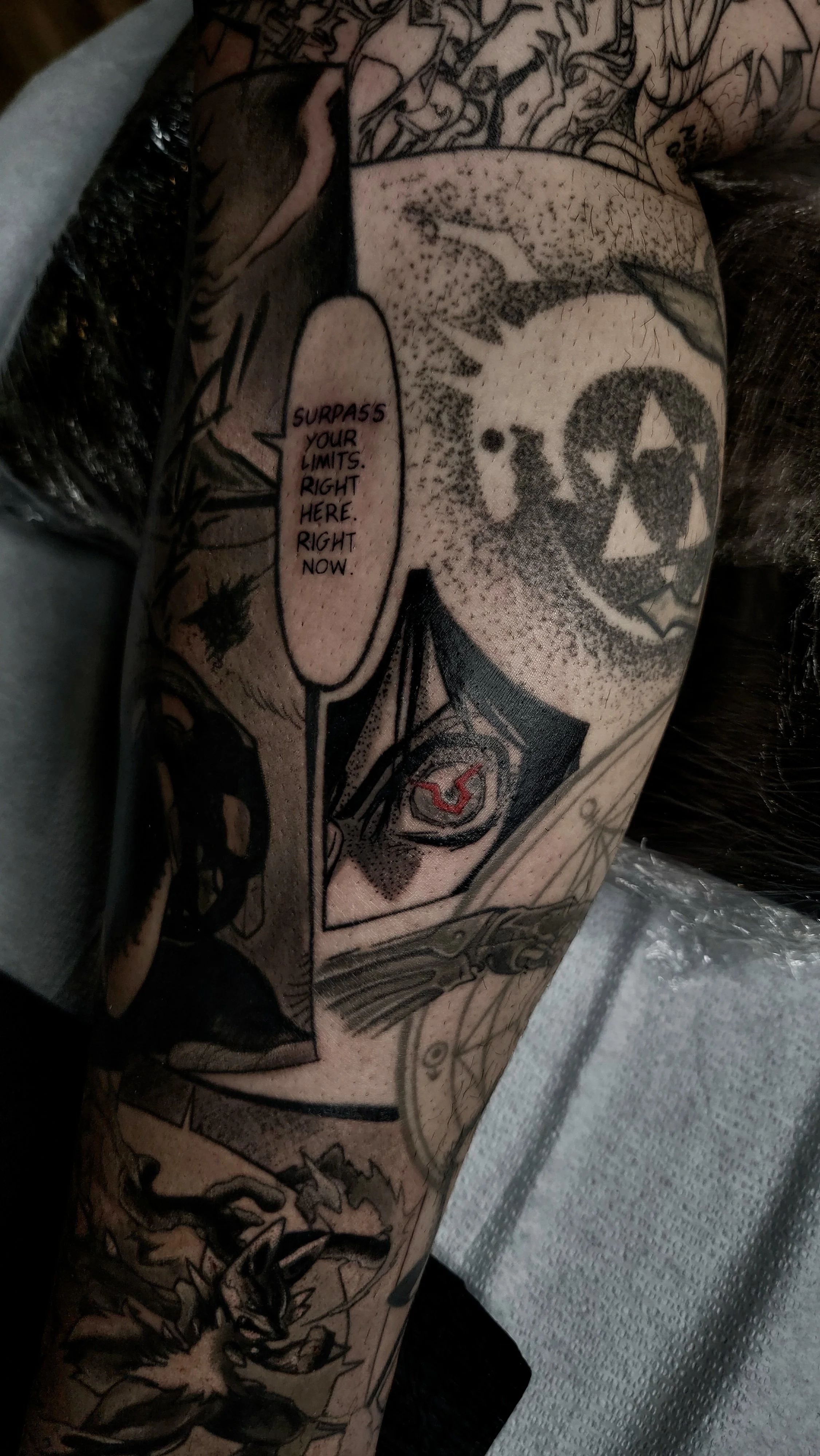 Close-up of a person's arm with a tattoo featuring various comic-style illustrations, including a speech bubble with the text "Surpass your limits right here right now," and a shield with a stylized symbol.