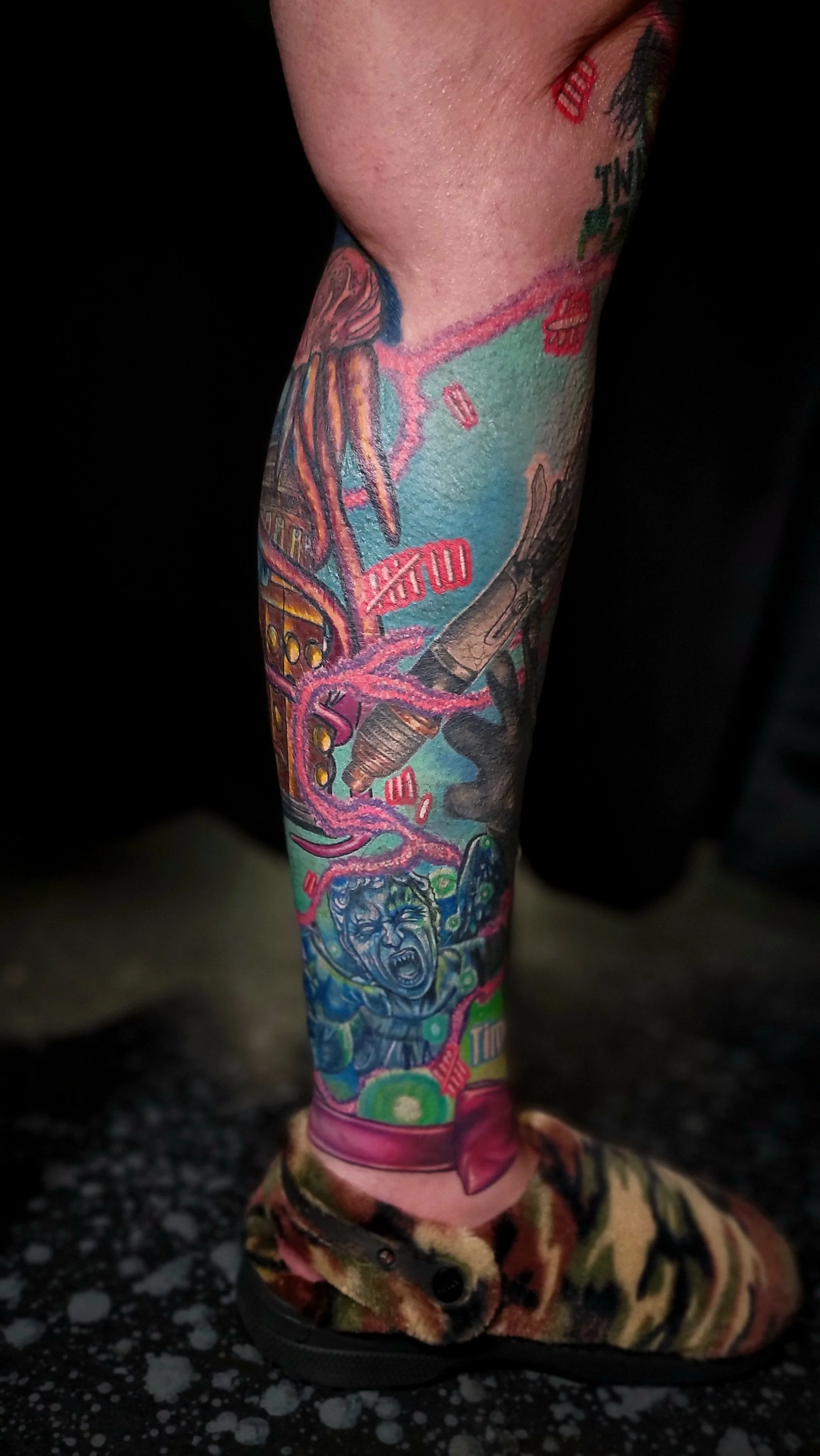 Close-up of a person's arm and leg tattooed with colorful, detailed artwork including a screaming face, a syringe, and other vivid designs, with the person wearing a camouflage-patterned shoe.