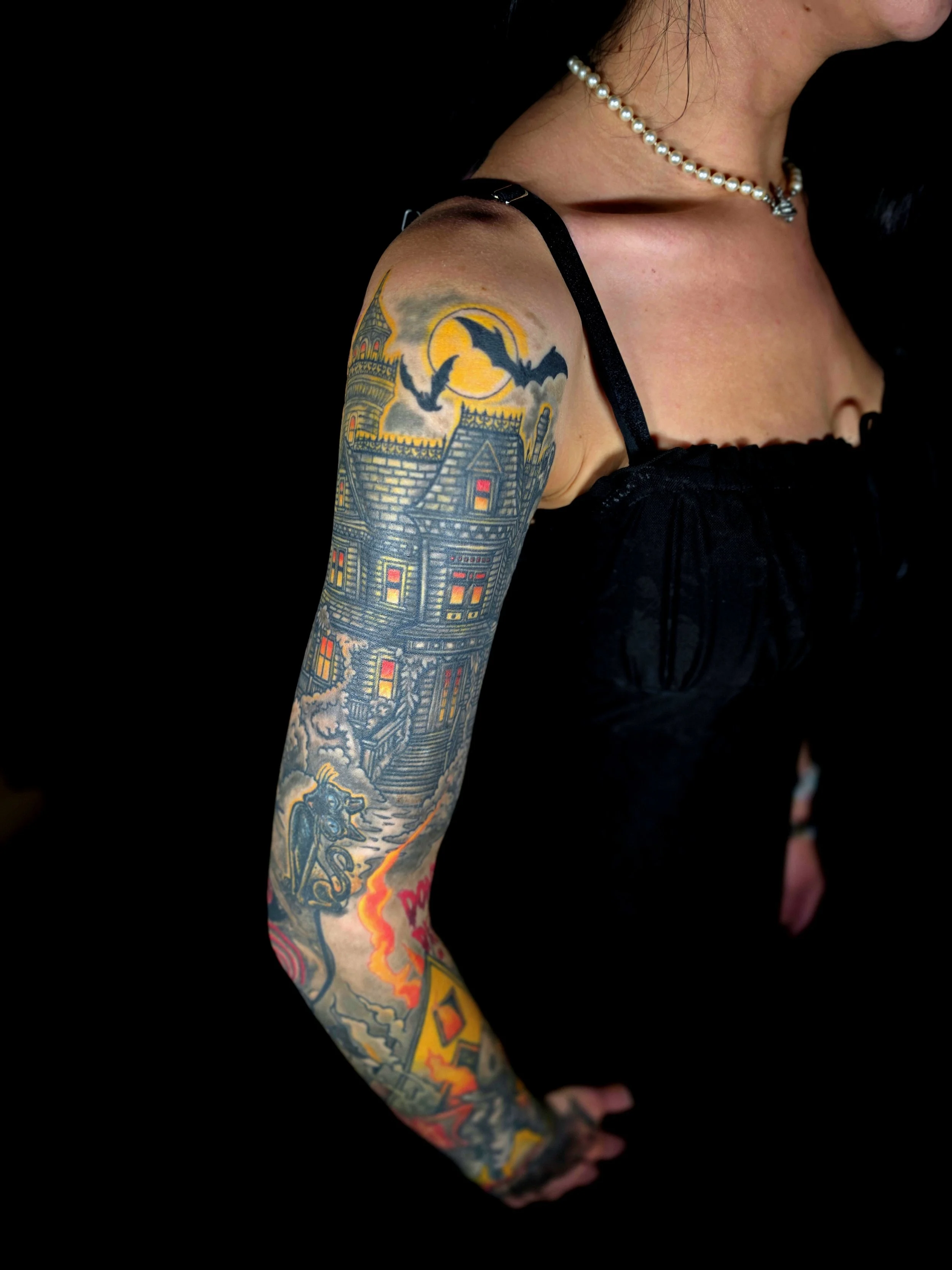 A person's arm with colorful tattoos, including a haunted house, a witch, and flames, against a black background. The person is wearing a pearl necklace and a black sleeveless top.