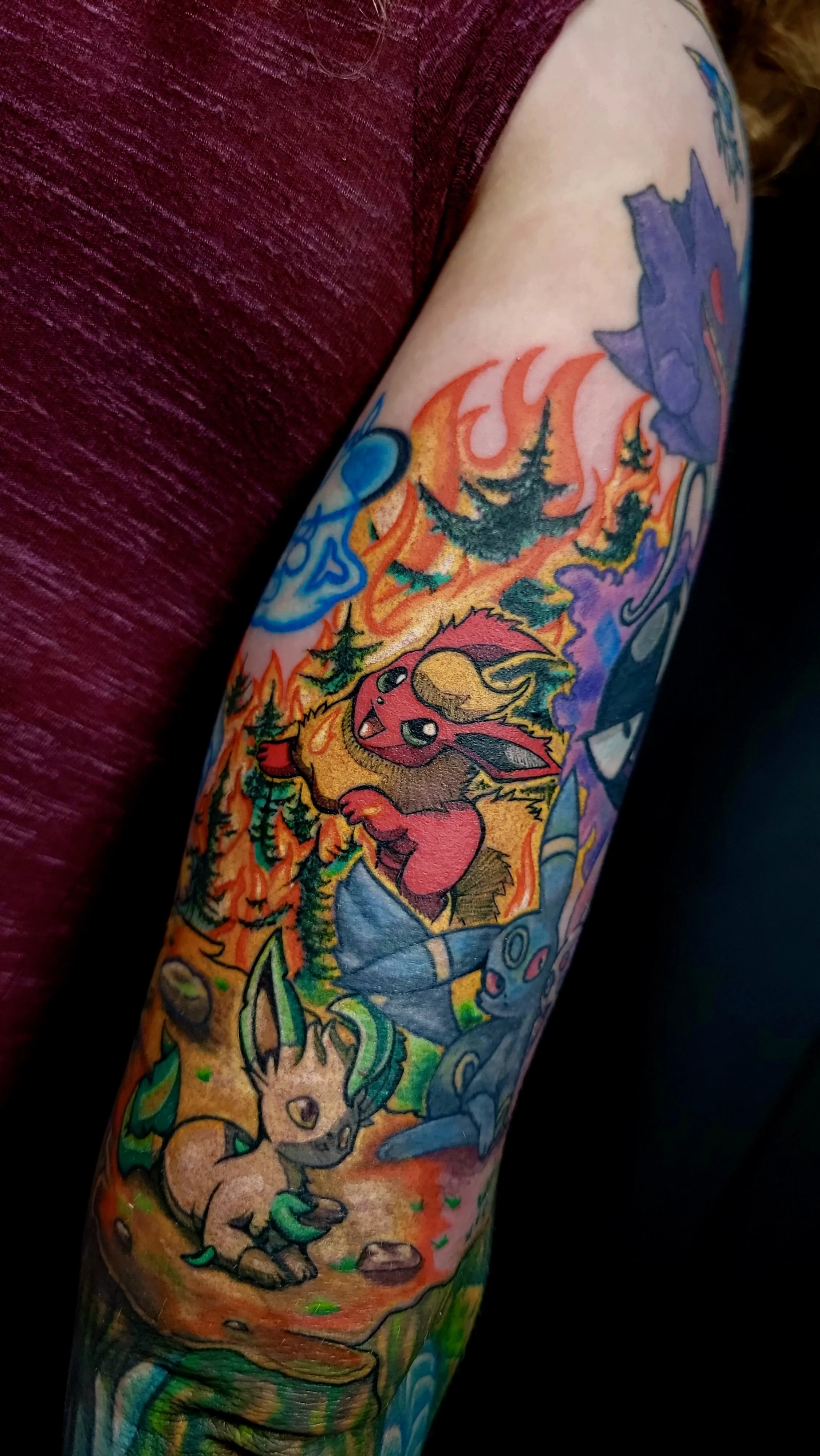 Close-up of a colorful tattoo on a person's arm featuring Pokémon characters including Eevee, Vaporeon, Flareon, and Jolteon, with fire and water elements in the background.