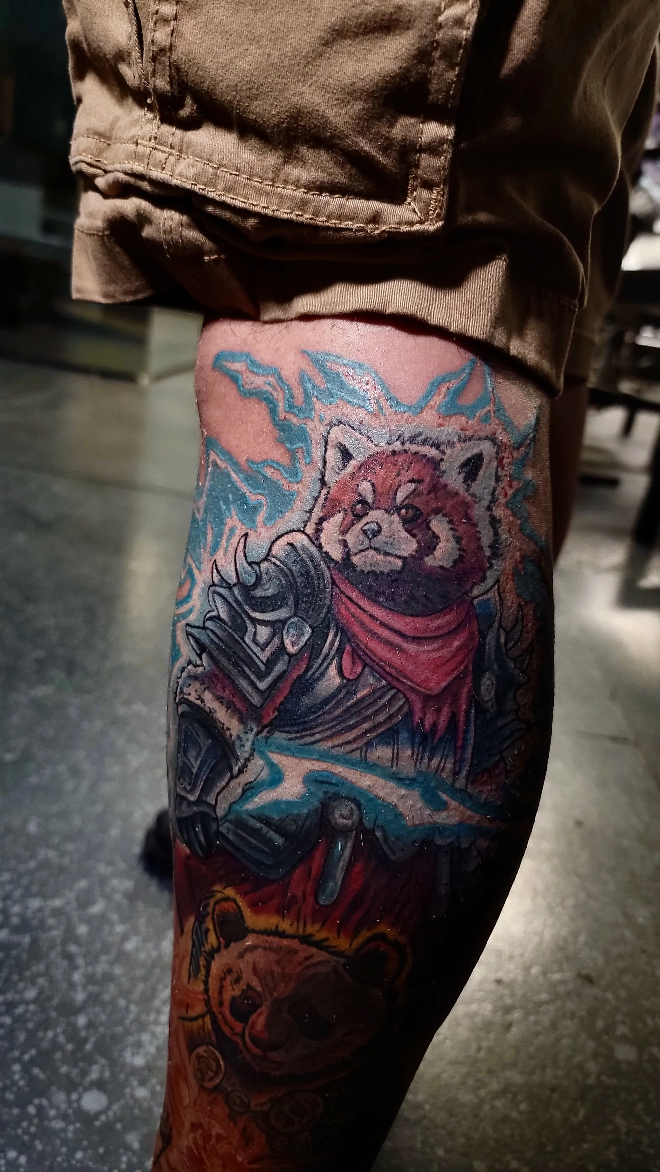 Tattoo of a red panda wearing armor, surrounded by colorful lightning and flames, on a person's leg.
