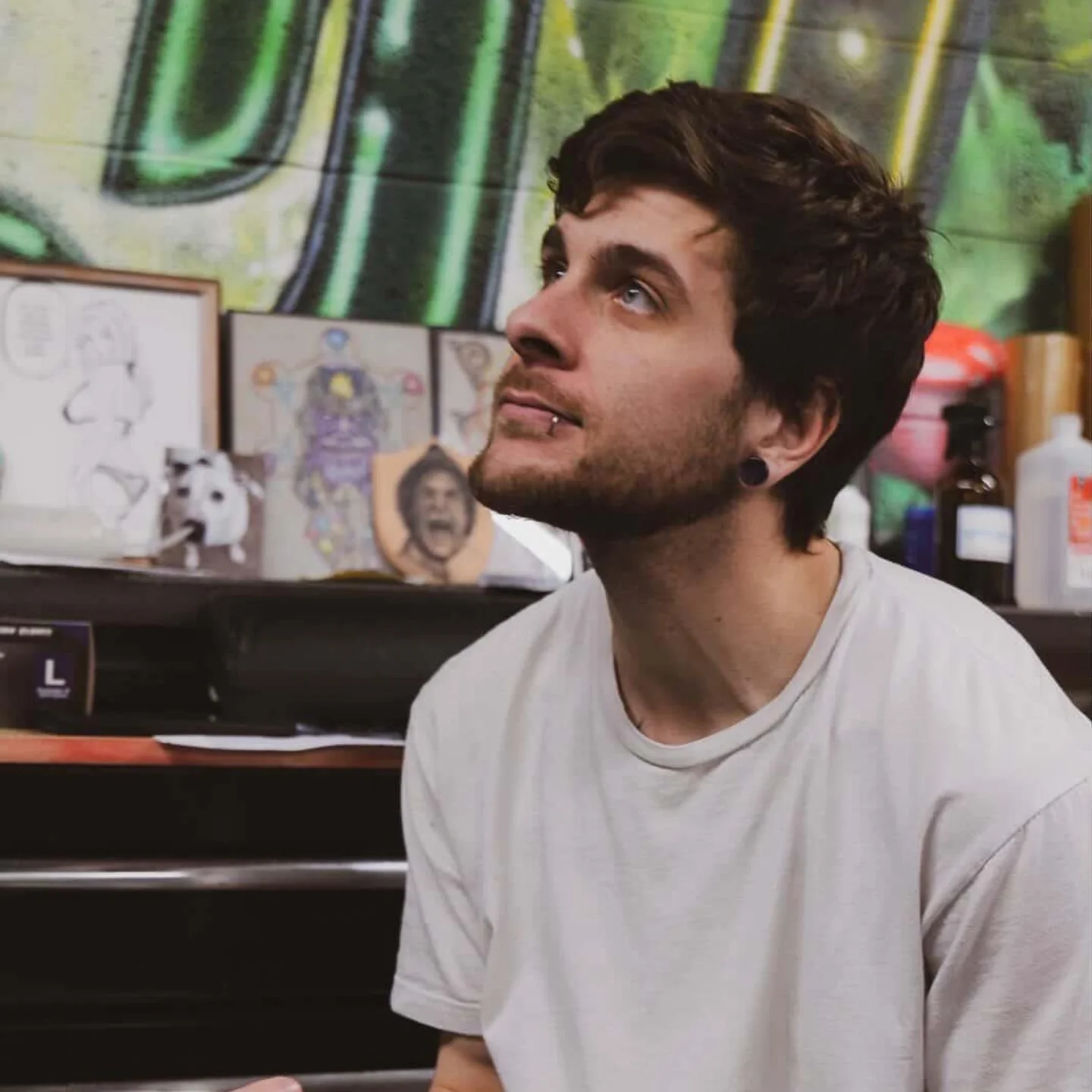 A young man with dark curly hair, pierced ears, and a lip piercing sitting in a tattoo studio with colorful artwork and tattoo supplies in the background.