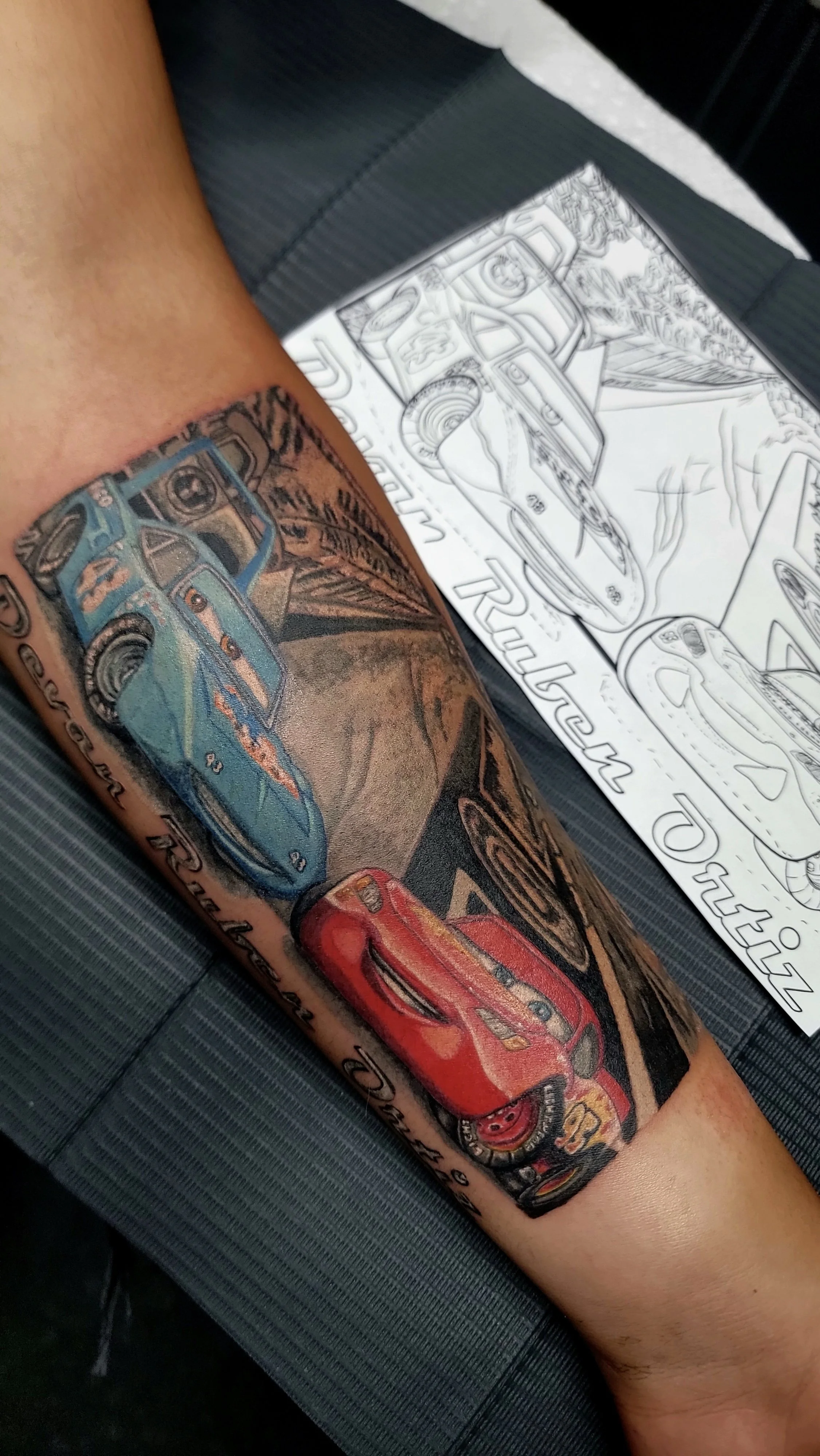 Colorful tattoo of three cars on a person's forearm, with a sketch of a similar car drawing on paper beside it.