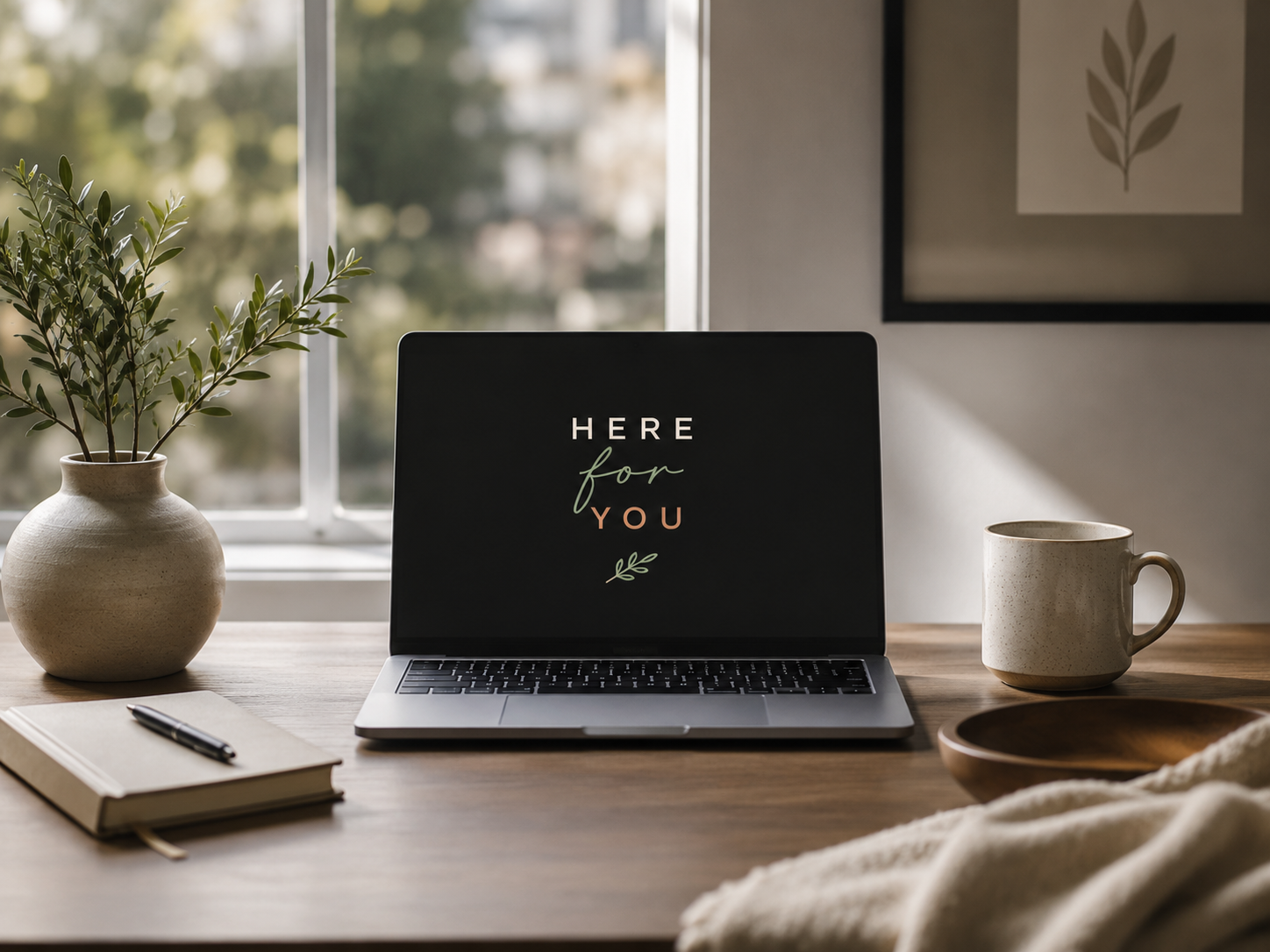 A laptop on a desk displaying 'Here for You' on the screen, with a potted plant, a notebook with a pen, a ceramic mug, and a wooden bowl nearby, in front of a window with sunlight streaming in.