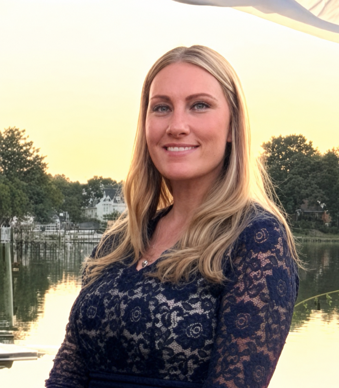 A woman with long blonde hair wearing a dark blue lace dress smiling outdoors near a body of water with trees and houses in the background during sunset.