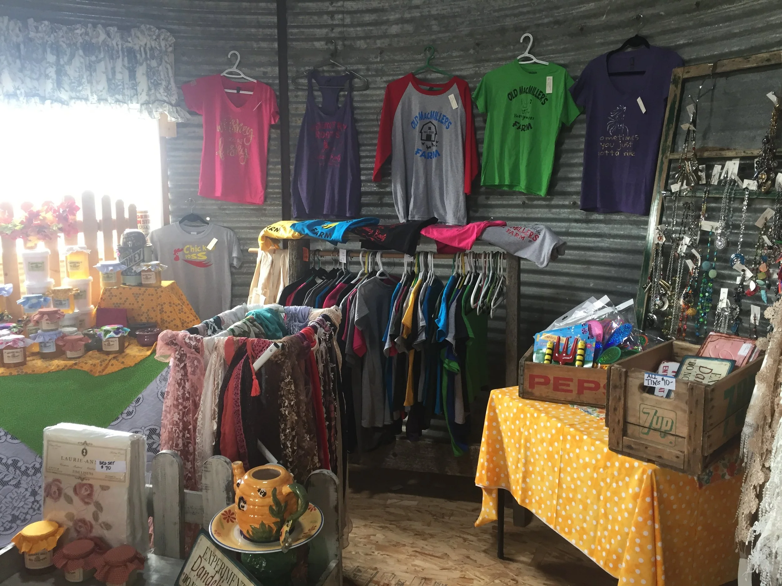 Display of various clothing items, jewelry, and home decor at a thrift or flea market stand, with shirts and dresses hanging on the wall and jewelry on a rack, along with a yellow polka dot tablecloth on a table.