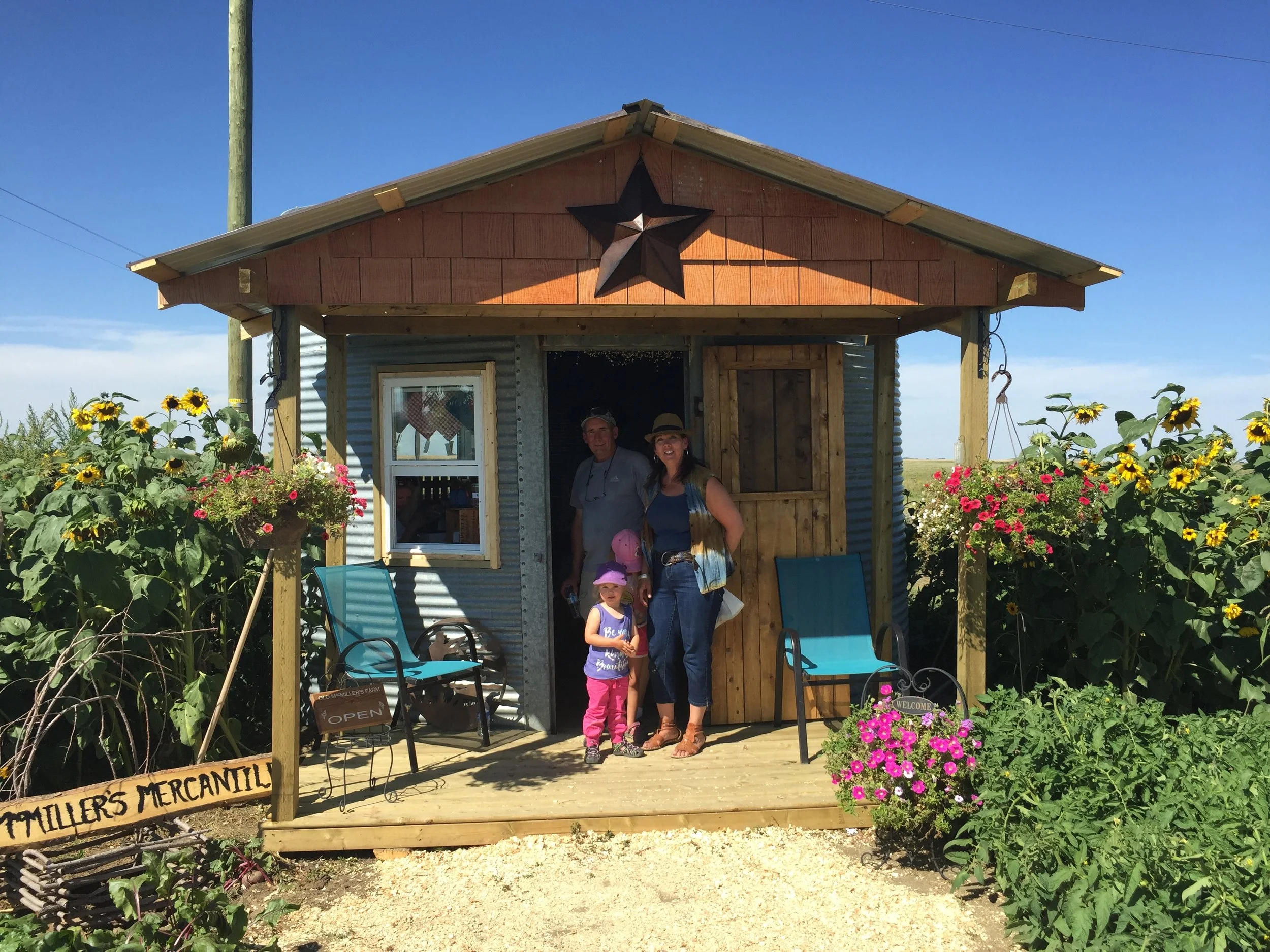 A small wooden market stand with a black star decoration on the front, surrounded by colorful flowers, with three people standing in front, including a woman, a man, and a young girl wearing a pink hat. The sign indicates it is a mercantile.