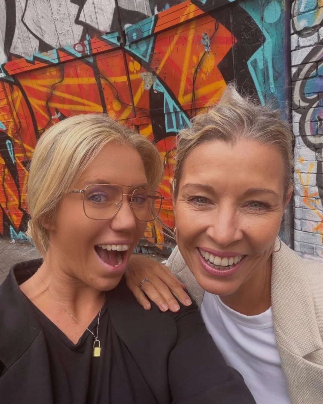 Two women smiling and taking a selfie in front of a vibrant graffiti wall featuring abstract designs in orange, blue, and yellow.