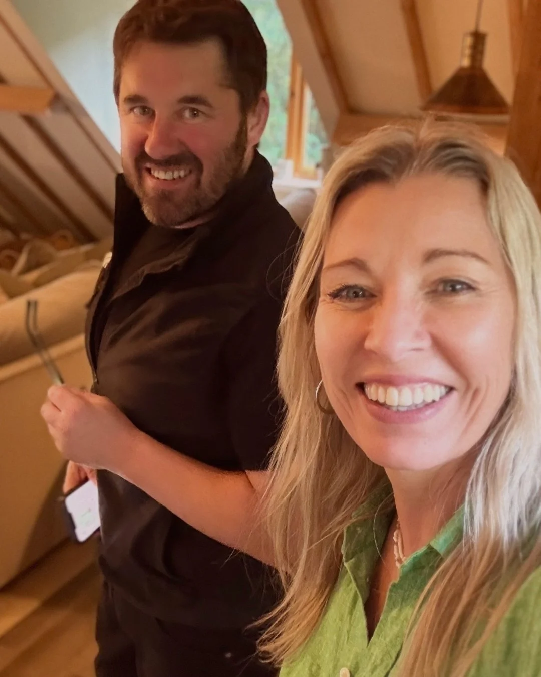 A smiling blonde woman in a green shirt taking a selfie with a smiling dark-haired man in a black shirt inside a wooden cabin.