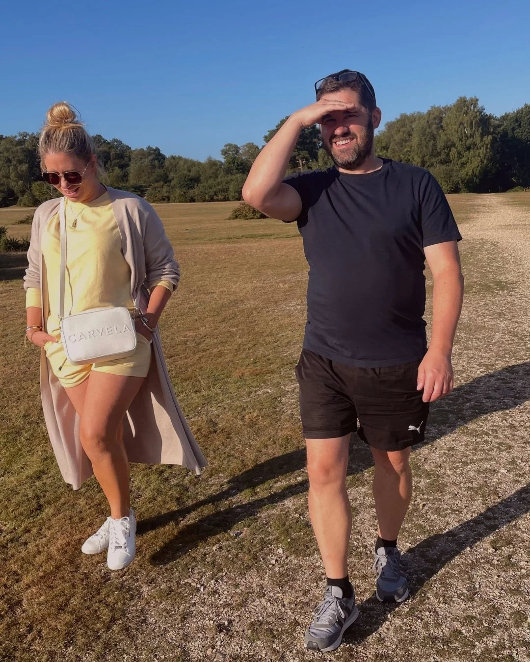 A man and woman outdoors on a sunny day, standing on a grassy field with trees in the background. The woman is wearing a light yellow outfit with sneakers, sunglasses, and a beige coat, while the man is wearing a black t-shirt, shorts, and sneakers, 