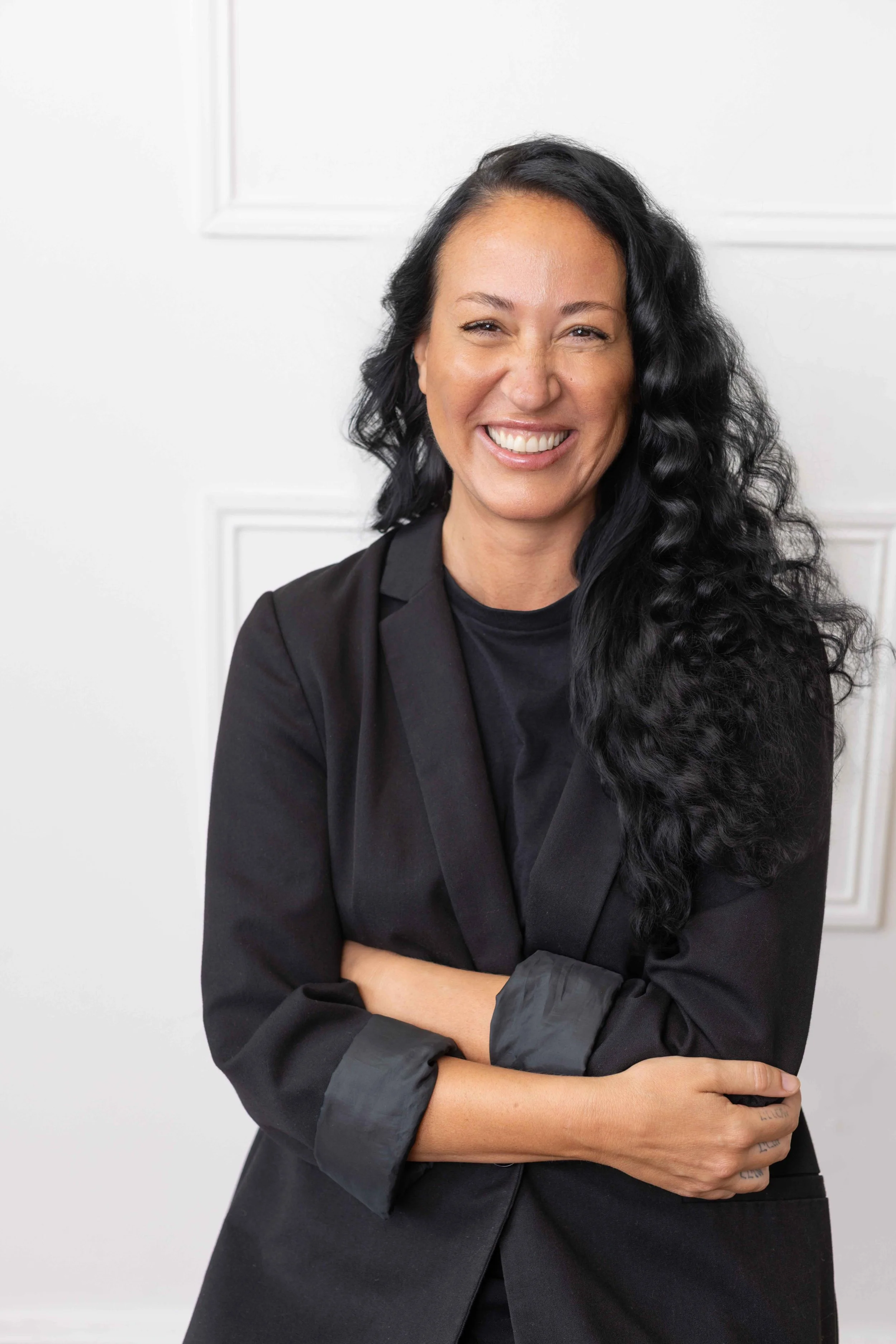 A woman with long, curly black hair and a wide smile, dressed in a black blazer and black shirt, standing in front of a white wall with decorative molding.