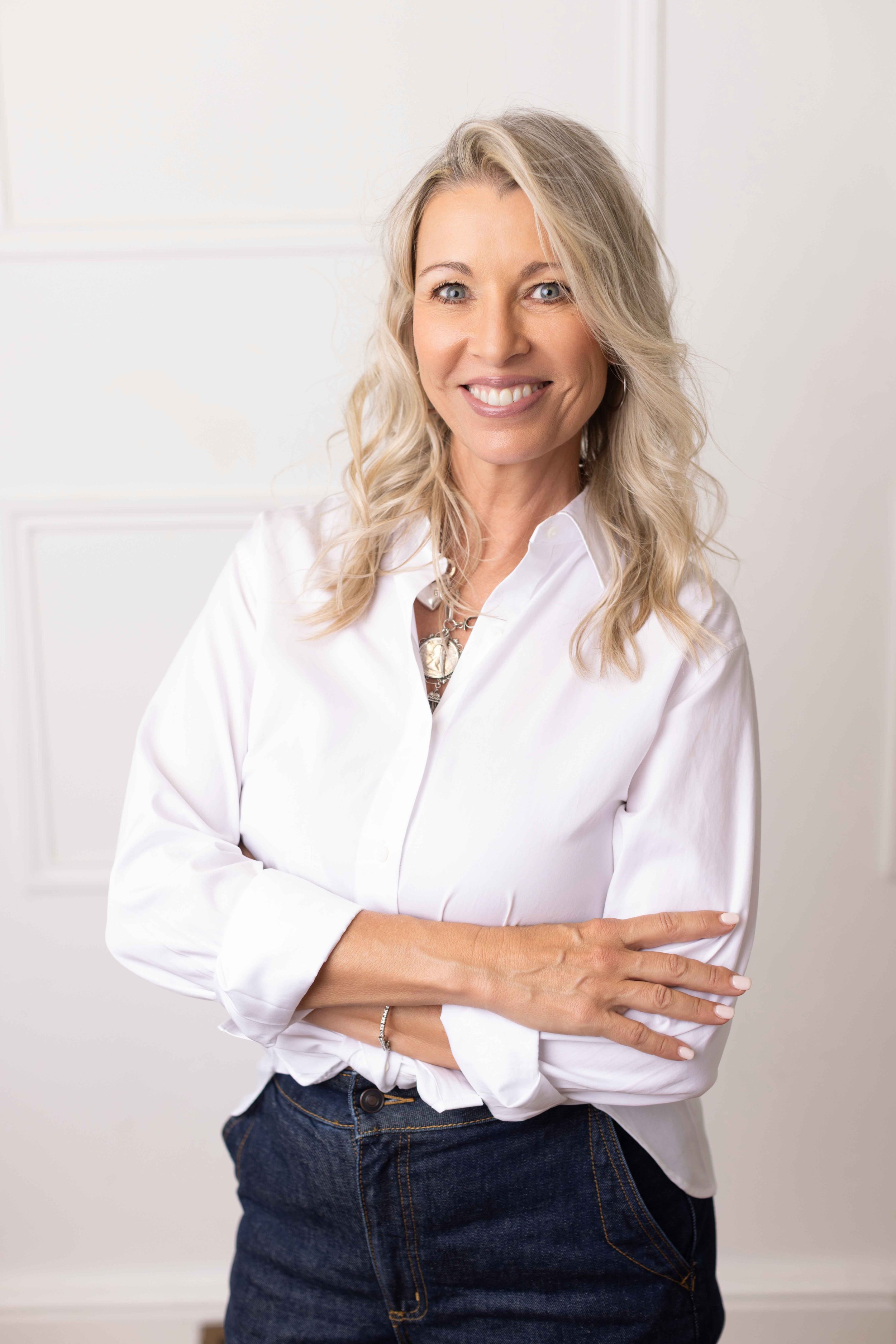 A woman with blonde, wavy hair smiling, wearing a white button-down shirt and dark jeans, her arms crossed in front of her.