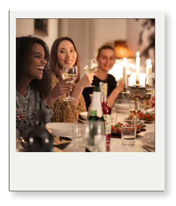 Three women at a dinner party raising glasses of white wine, smiling, with a table set with food, candles, and drinks, in a warmly lit room.