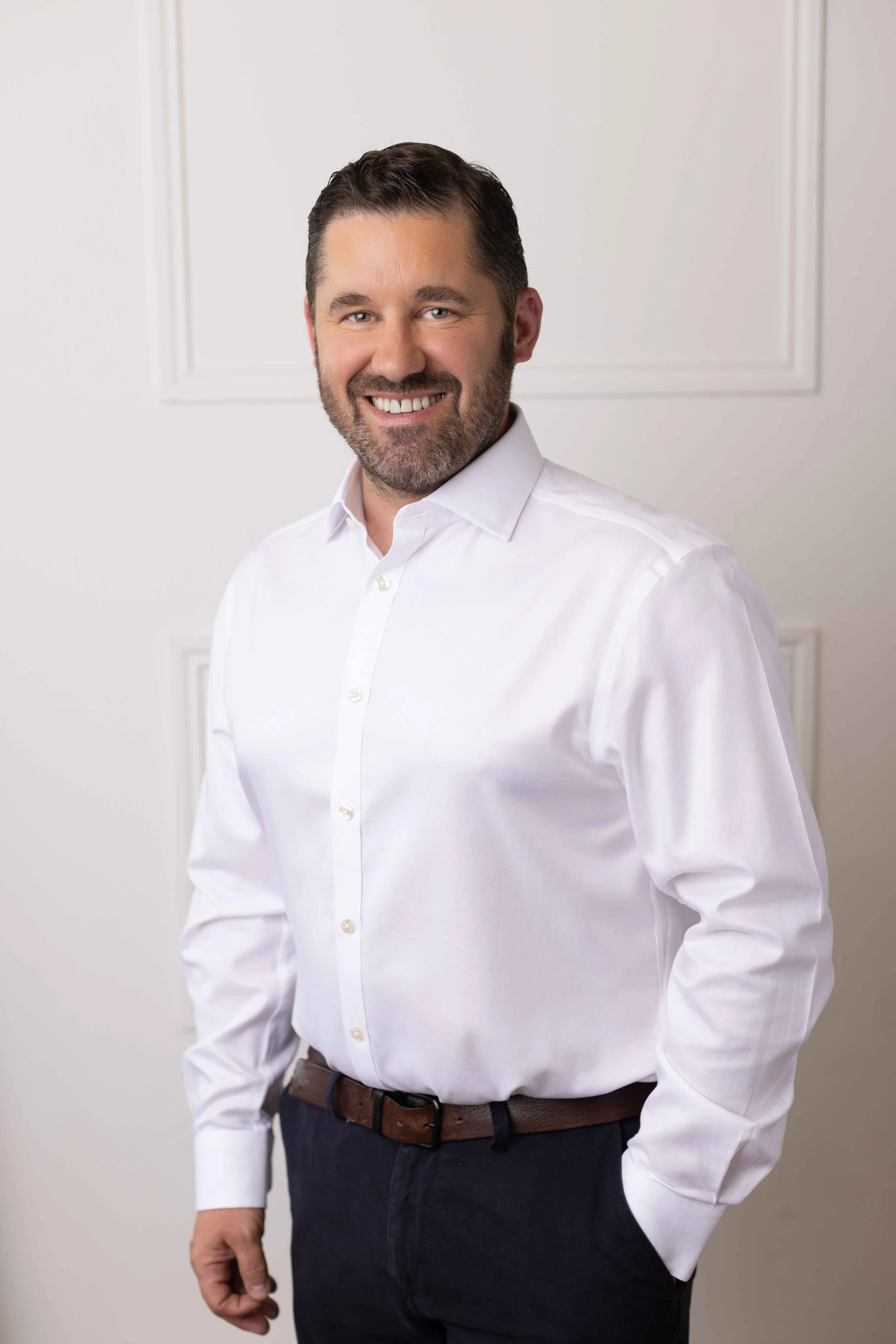 A man with dark hair and a beard, wearing a white dress shirt tucked into dark pants, standing against a plain white wall.