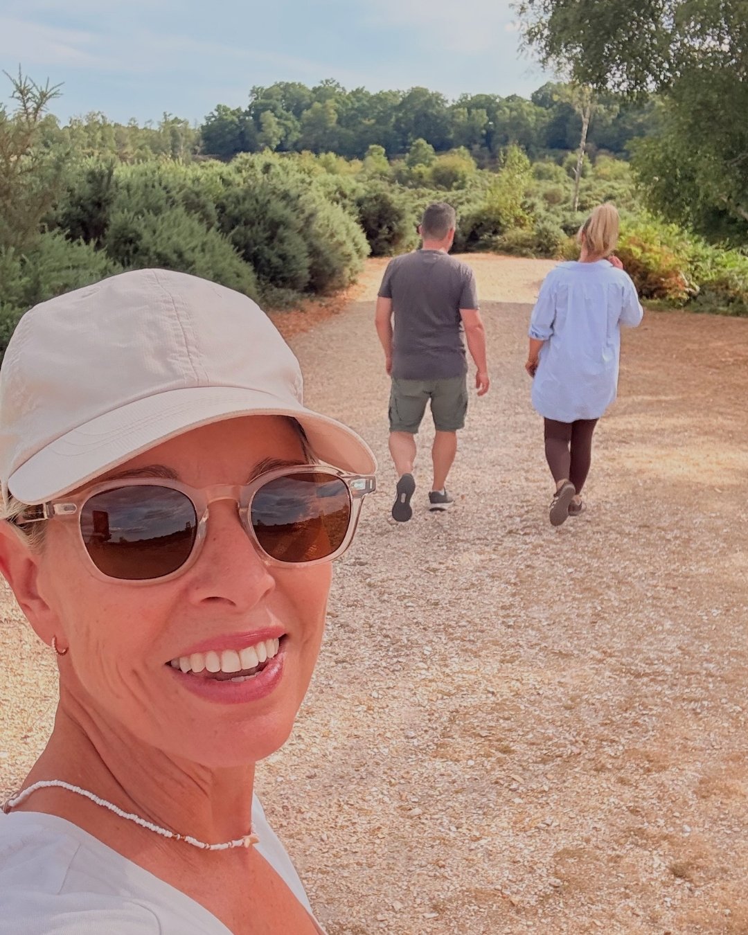Woman smiling wearing sunglasses, white cap, and a necklace, taking a selfie on a dirt trail with two people walking ahead in a nature setting.