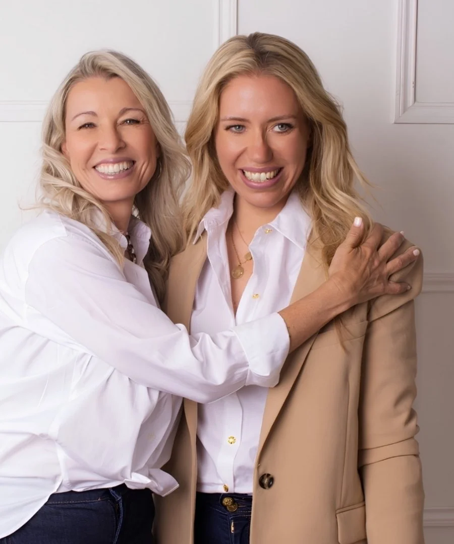 Two women smiling and embracing each other indoors, one in a white shirt and the other in a beige blazer and white shirt.