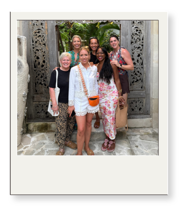 Six women smiling and posing for a photo in front of an ornate wooden gate with tropical greenery in the background.