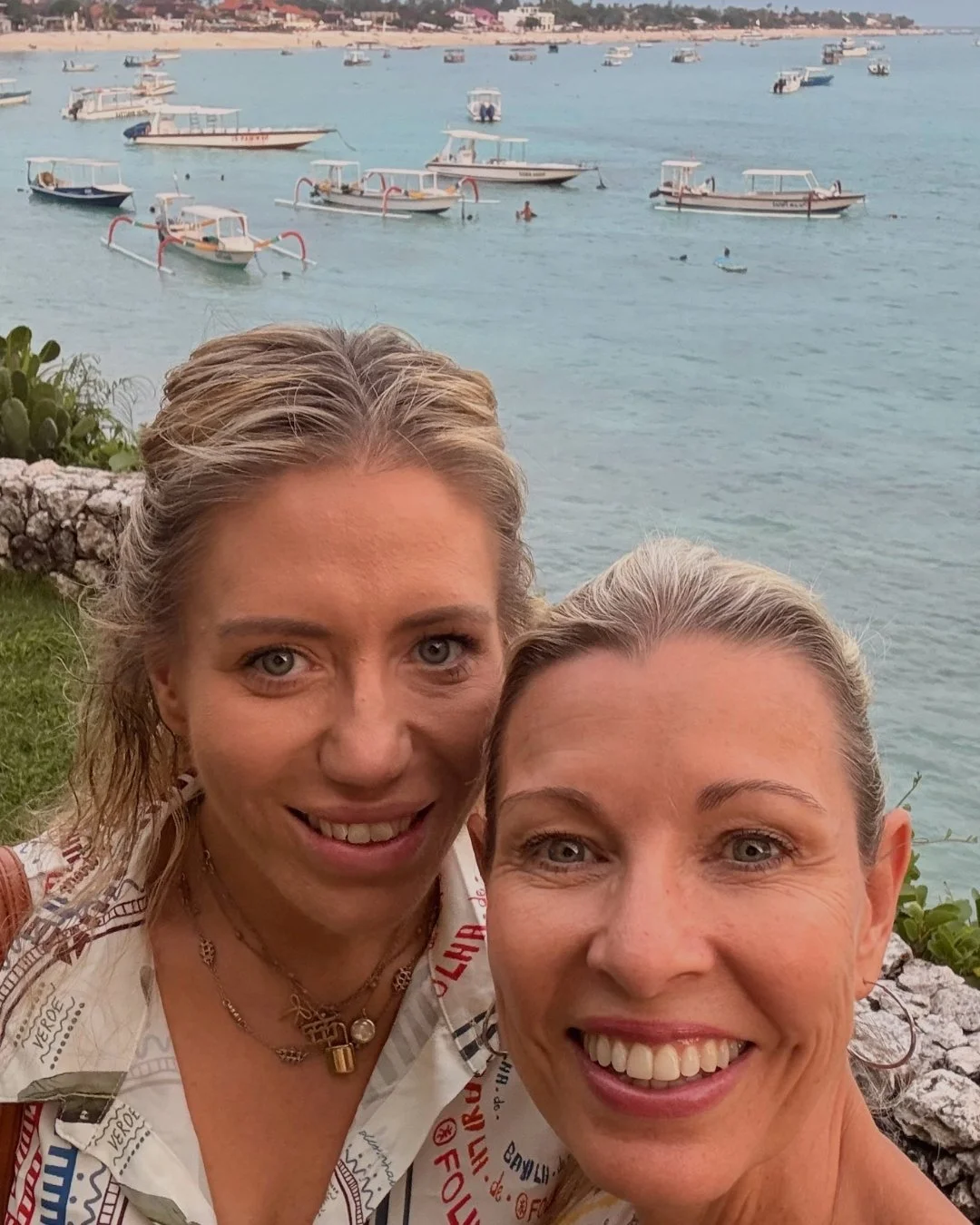 Two women smiling for a selfie near the water, with numerous boats anchored in the background.