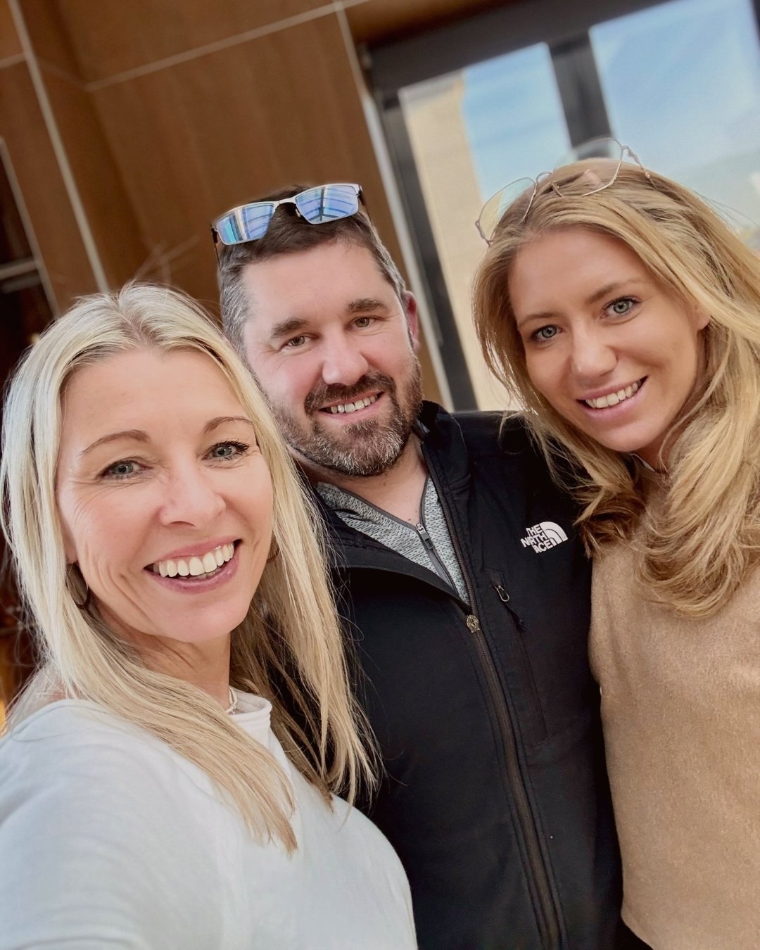Three people smiling for a selfie inside a room with large windows and wooden walls.