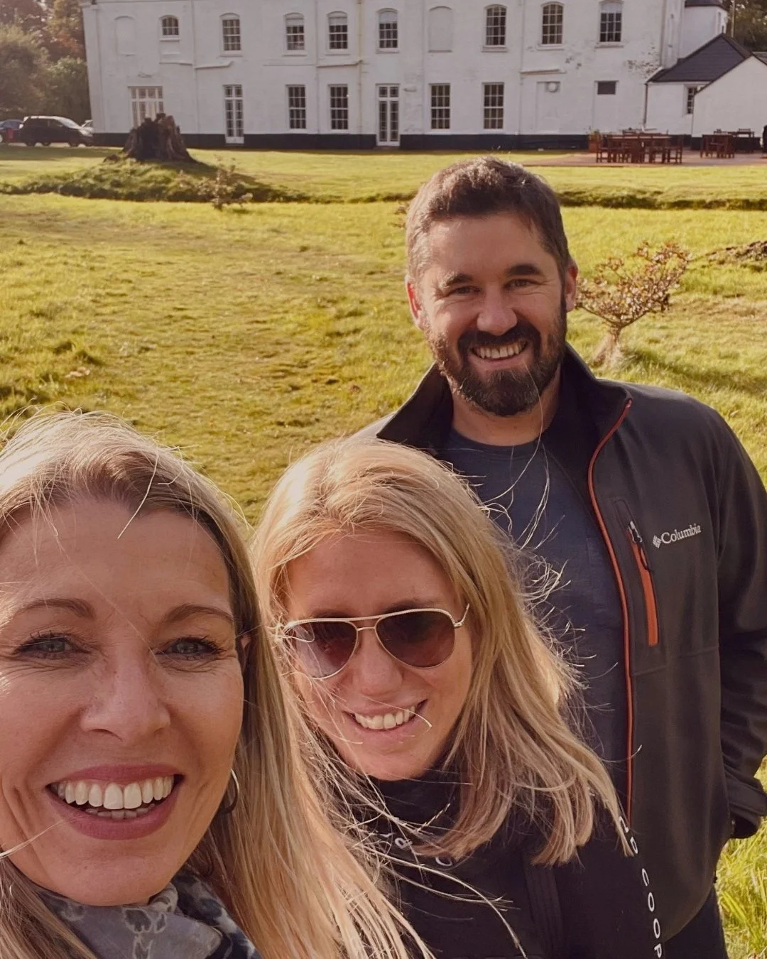 Three smiling people taking a selfie outdoors in front of a large white building with a grassy lawn.
