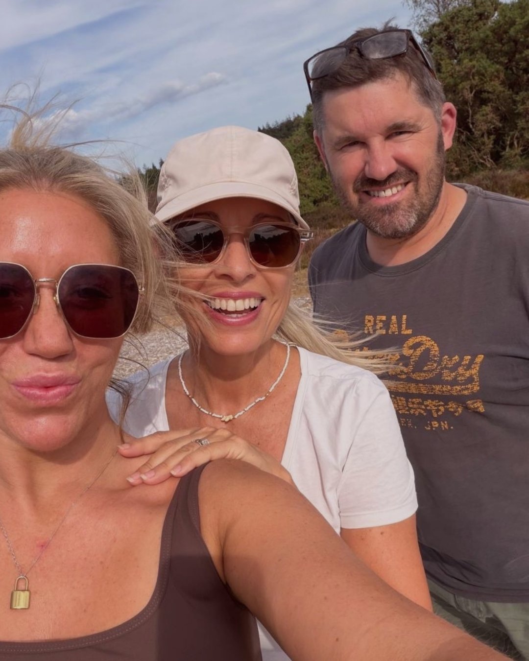 Three smiling adults taking a selfie outdoors with a cloudy sky and trees in the background.