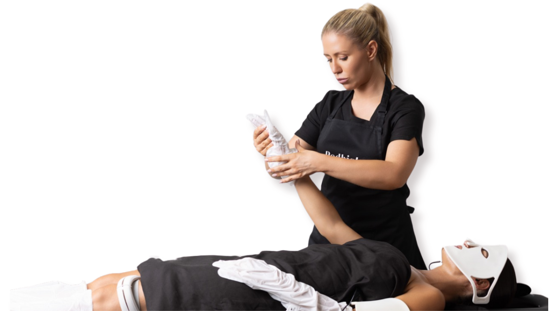 A woman in black scrubs puts on gloves while preparing a medical procedure on a person lying on a table, who is wearing a white mask and has a medical device on their leg.