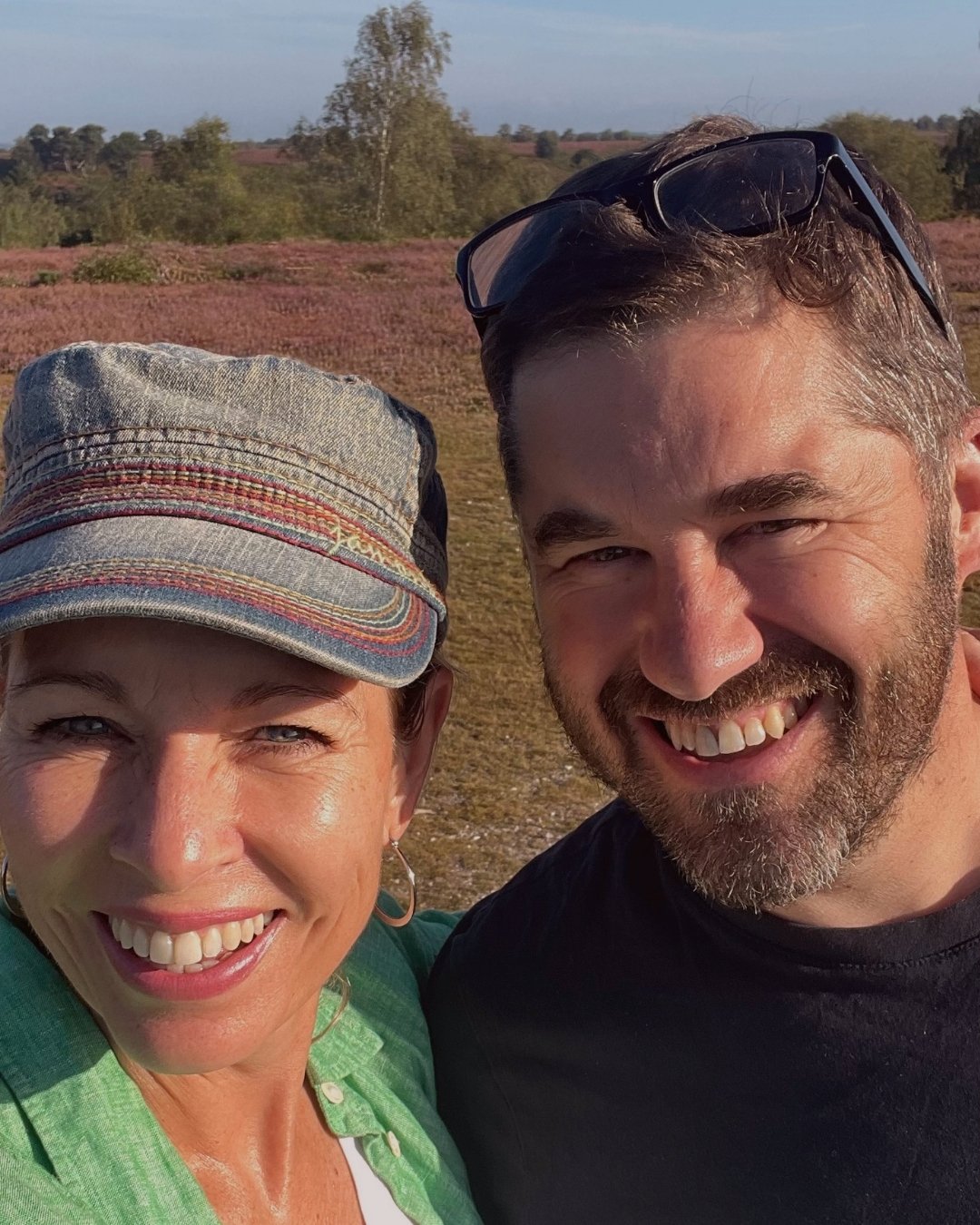 A smiling woman wearing a denim cap and a man with sunglasses on his head, together outdoors in a field with pinkish-purple vegetation and trees in the background.