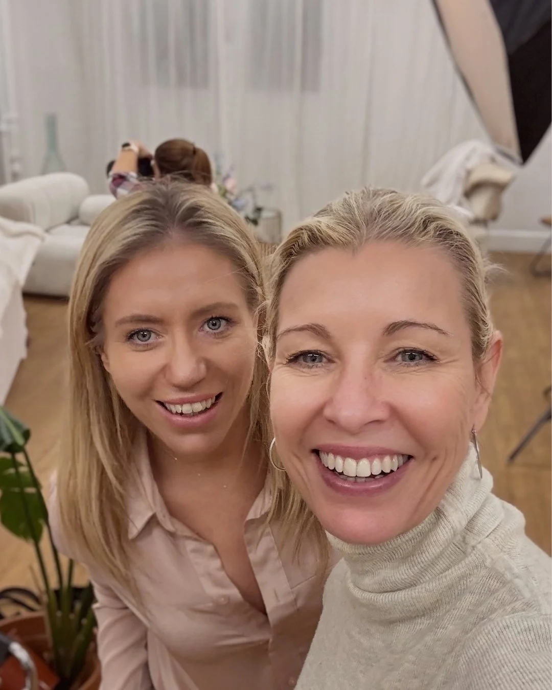 Two women smiling and taking a selfie in a living room with a person in the background doing makeup or hair.
