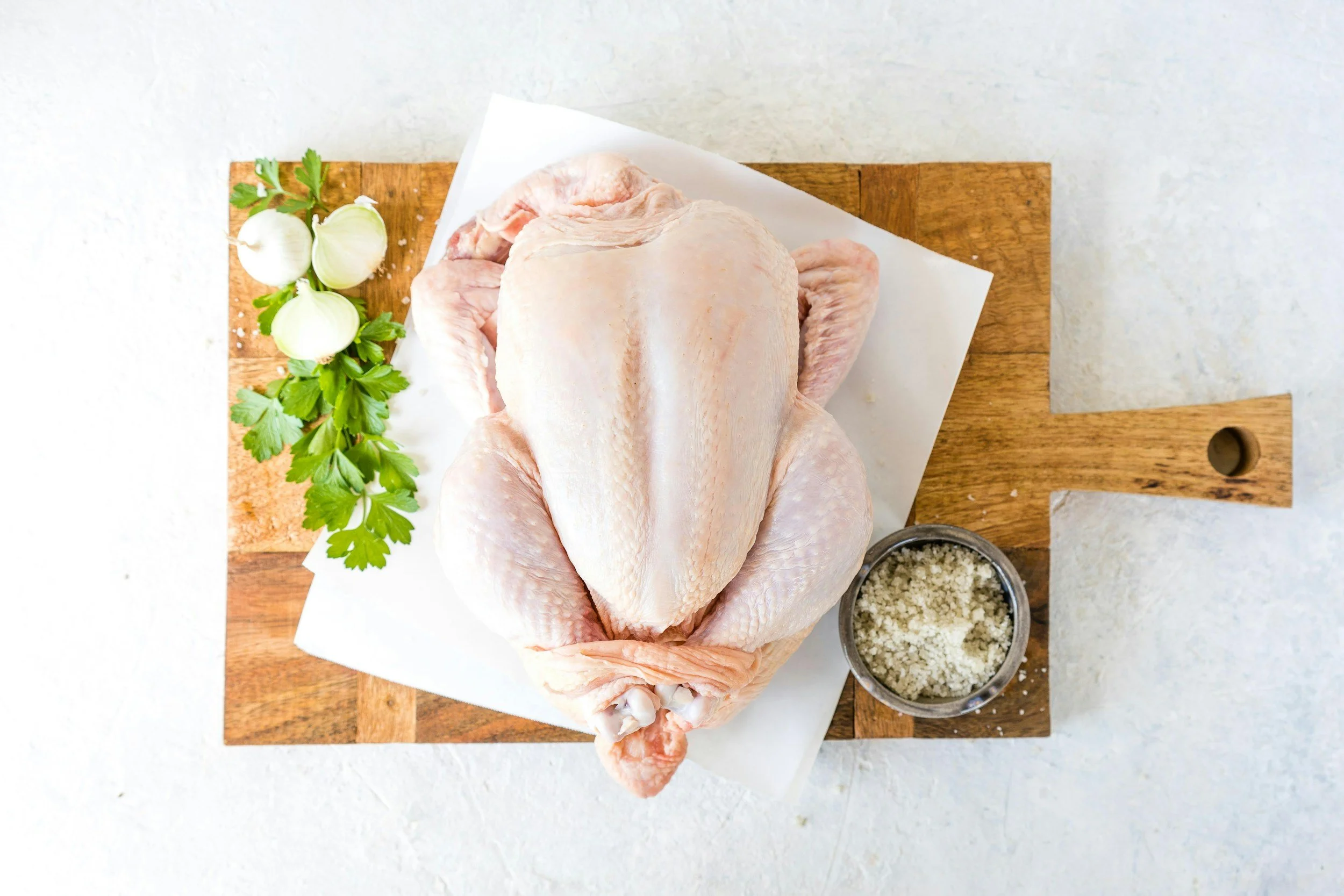 Raw whole chicken on a cutting board with garlic, parsley, and bowls of seasonings.