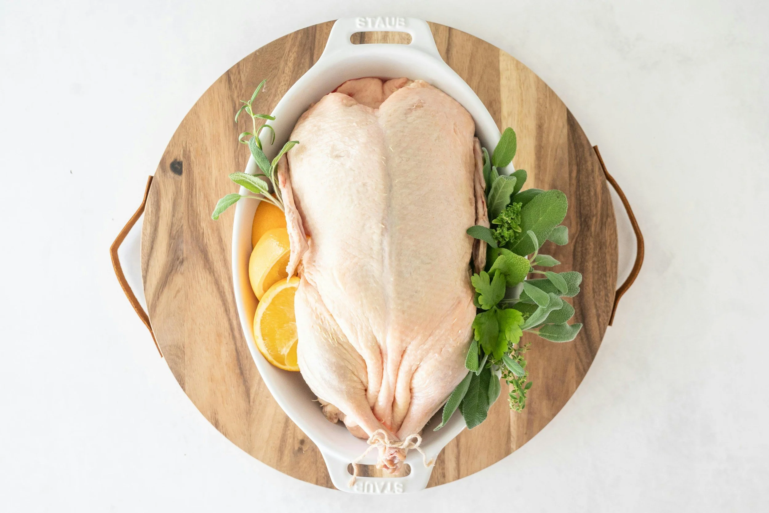 Raw duck in a white oval dish with lemon slices and fresh herbs, on a round wooden cutting board.