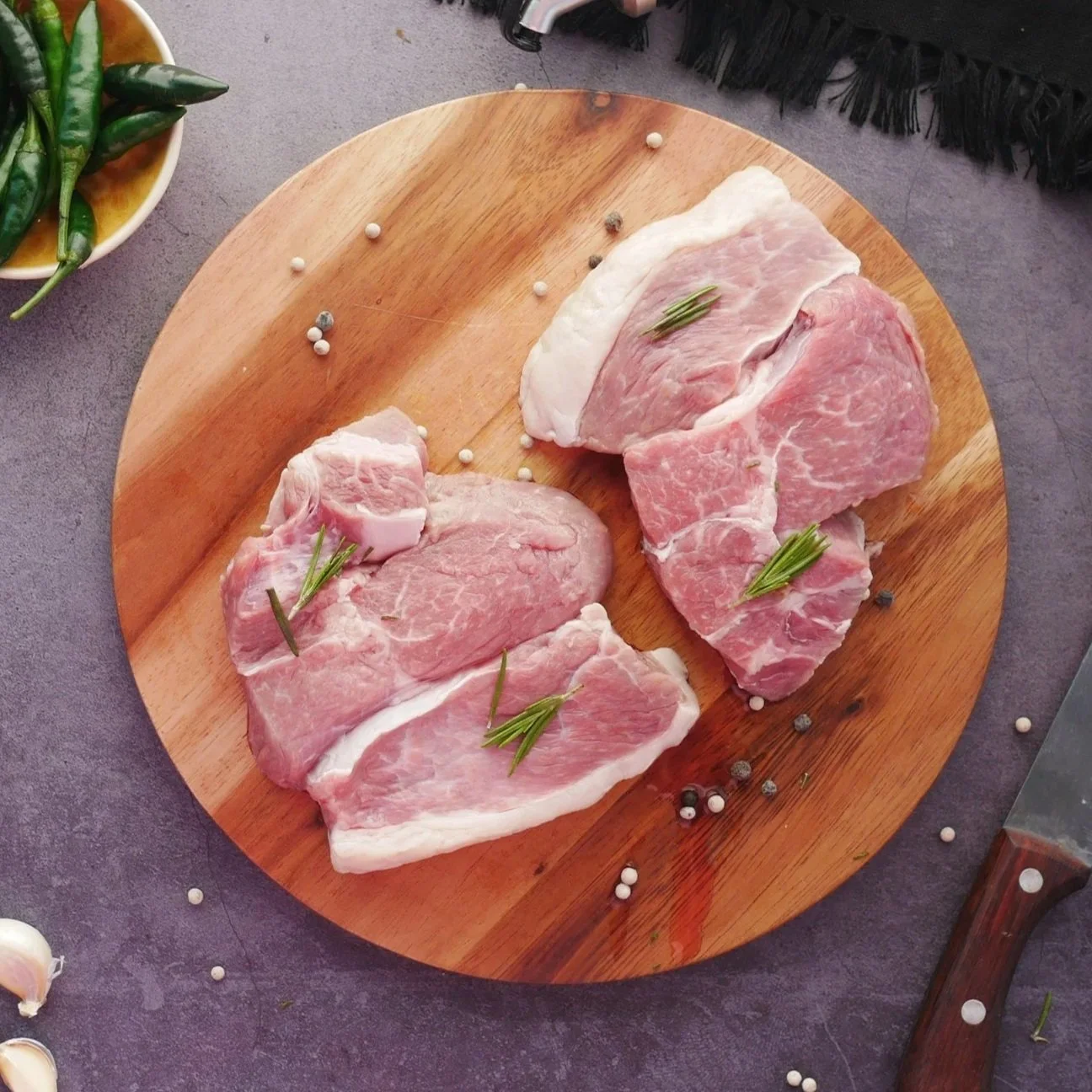 Raw pork chops with rosemary on a round wooden cutting board, garnished with peppercorns and placed on a gray surface with a bowl of green chili peppers on the side.