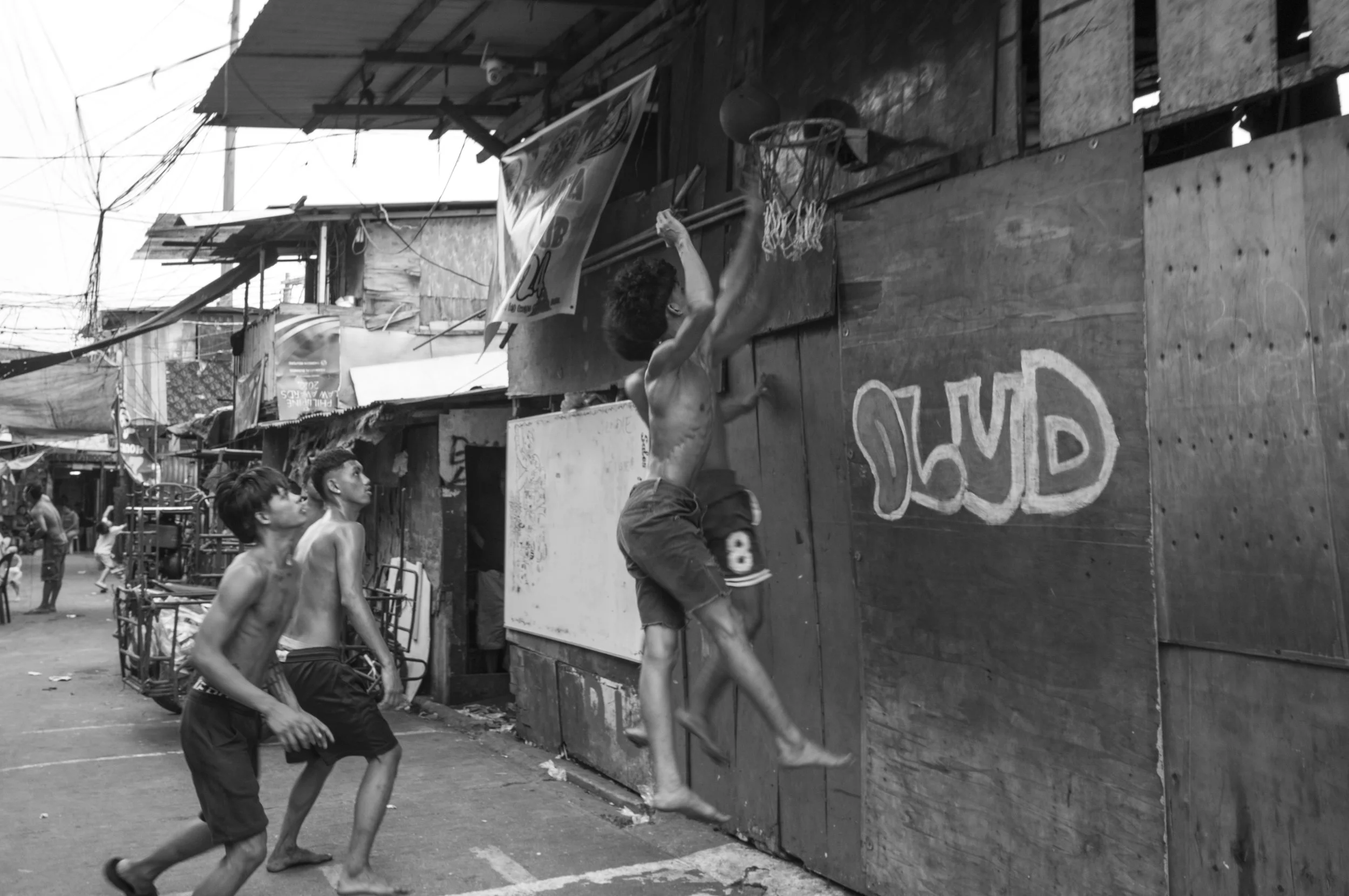 Boys playing basketball outdoors on a makeshift court, jumping to make a shot at the hoop attached to a wooden wall, while other children watch nearby in a neighborhood setting with houses and utility wires in Tondo, Manila ©R. Hartel for odoVita.