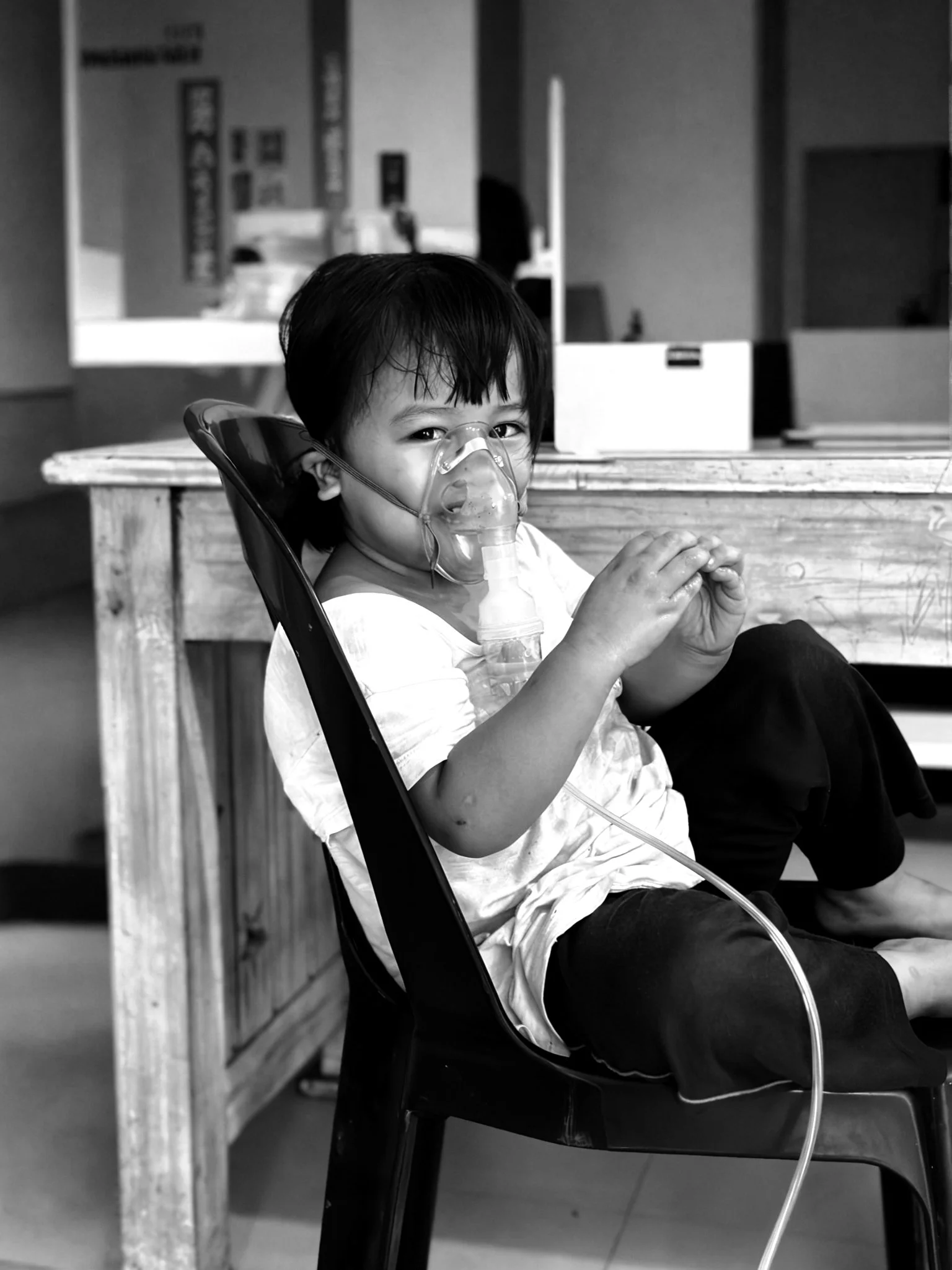 A young child with dark hair and a nasal oxygen mask, sitting on a black chair in a room, with his hands clasped. In the background, there is a wooden table, and a blurred figure is visible through a window or mirror in Tondo, Manila ©odoVita.