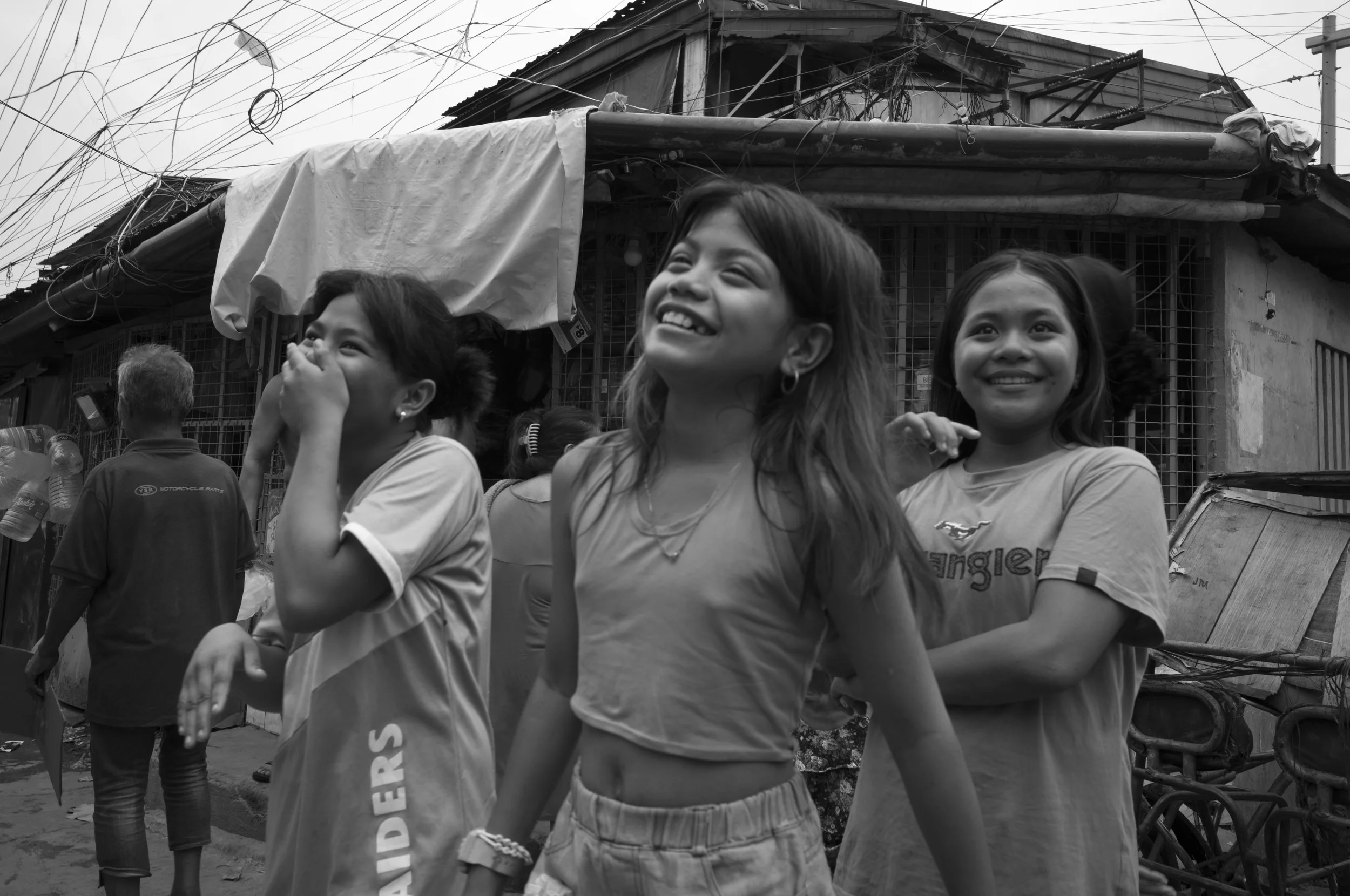 A black and white photo of three young girls smiling and walking in a street with a weathered building and electric wires overhead in Tondo, Manila ©R. Hartel for odoVita.