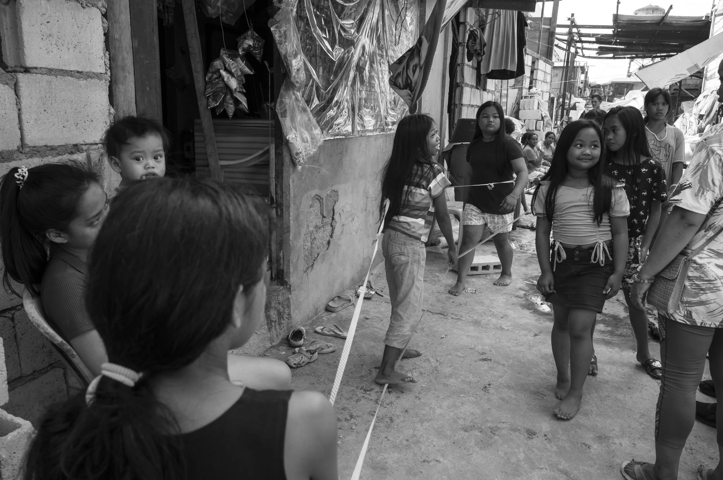 A group of children and young women standing outdoors in a row, with some smiling and others looking serious, in a community setting with makeshift structures and hanging clothes visible in the background in Tondo, Manila ©R. Hartel for odoVita.