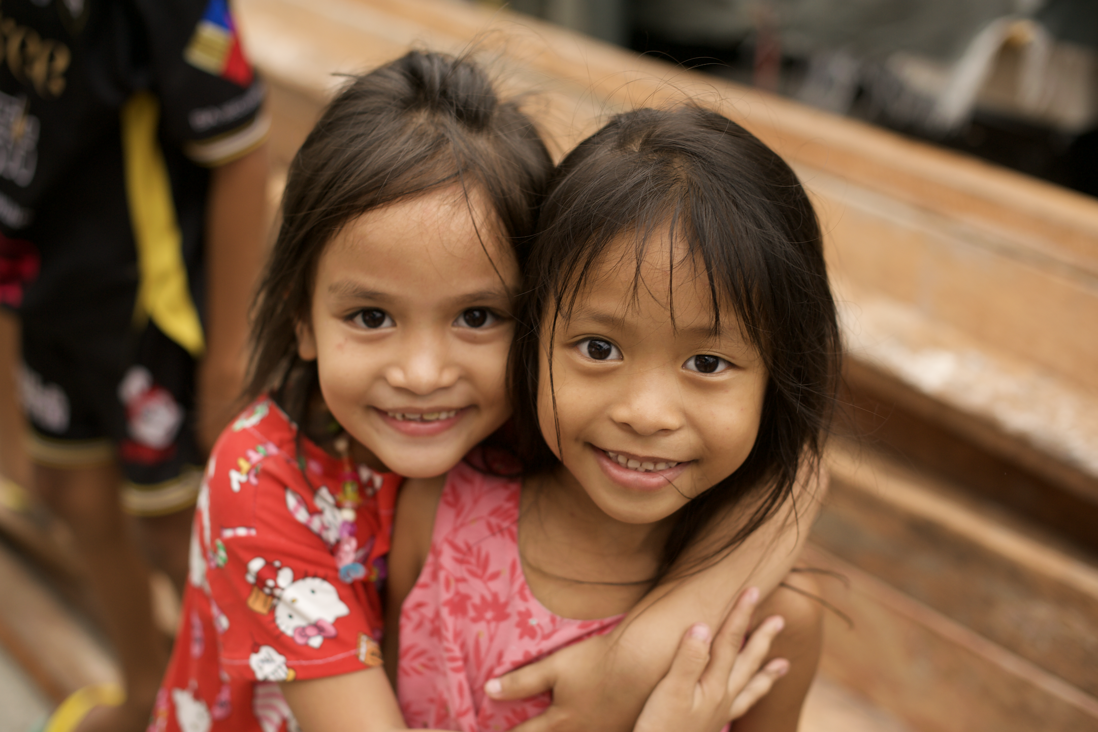 Two young girls smiling and hugging each other, with blurred children in colorful clothing in the background in Tondo, Manila ©R. Hartel for odoVita.
