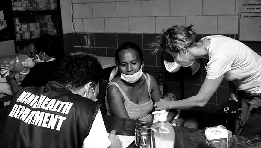 Health worker examining a woman holding a baby in a health clinic, with a health department worker and another woman in the background in Tondo, Manila ©odoVita.