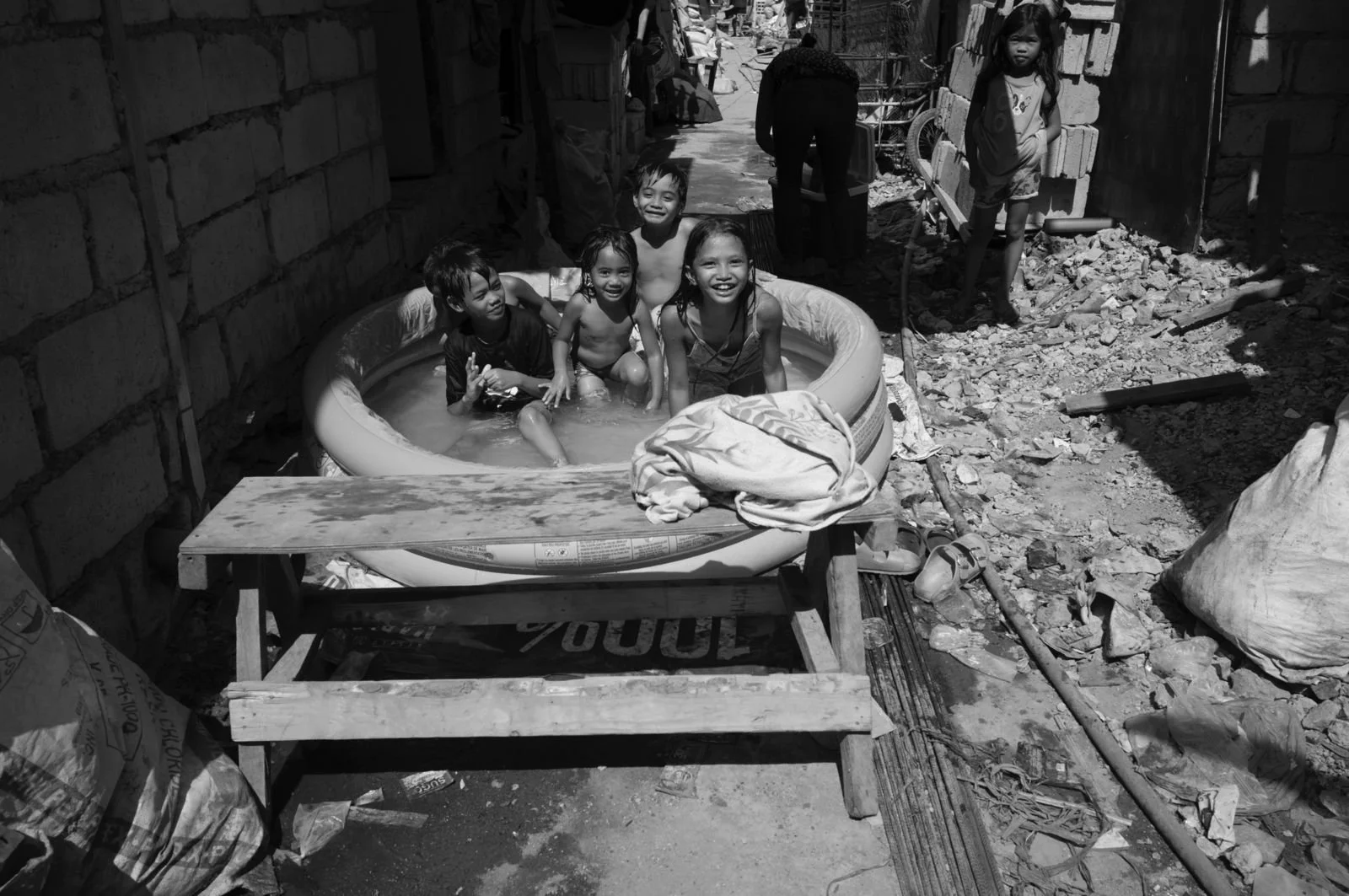 Five children are playing and smiling in a small inflatable swimming pool in an outdoor, makeshift area surrounded by debris and construction materials. The scene is in black and white in Tondo, Manila ©R. Hartel for odoVita.