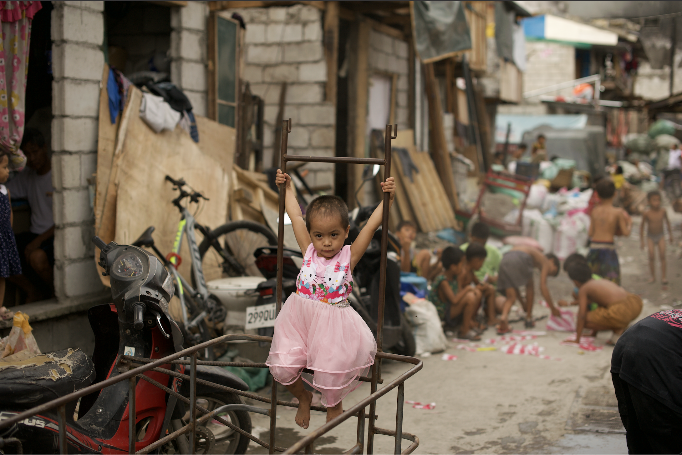 A young girl dressed as a princess standing inside a metal cart with other people and makeshift houses in the background of a crowded outdoor area in Tondo, Manila. ©odoVita