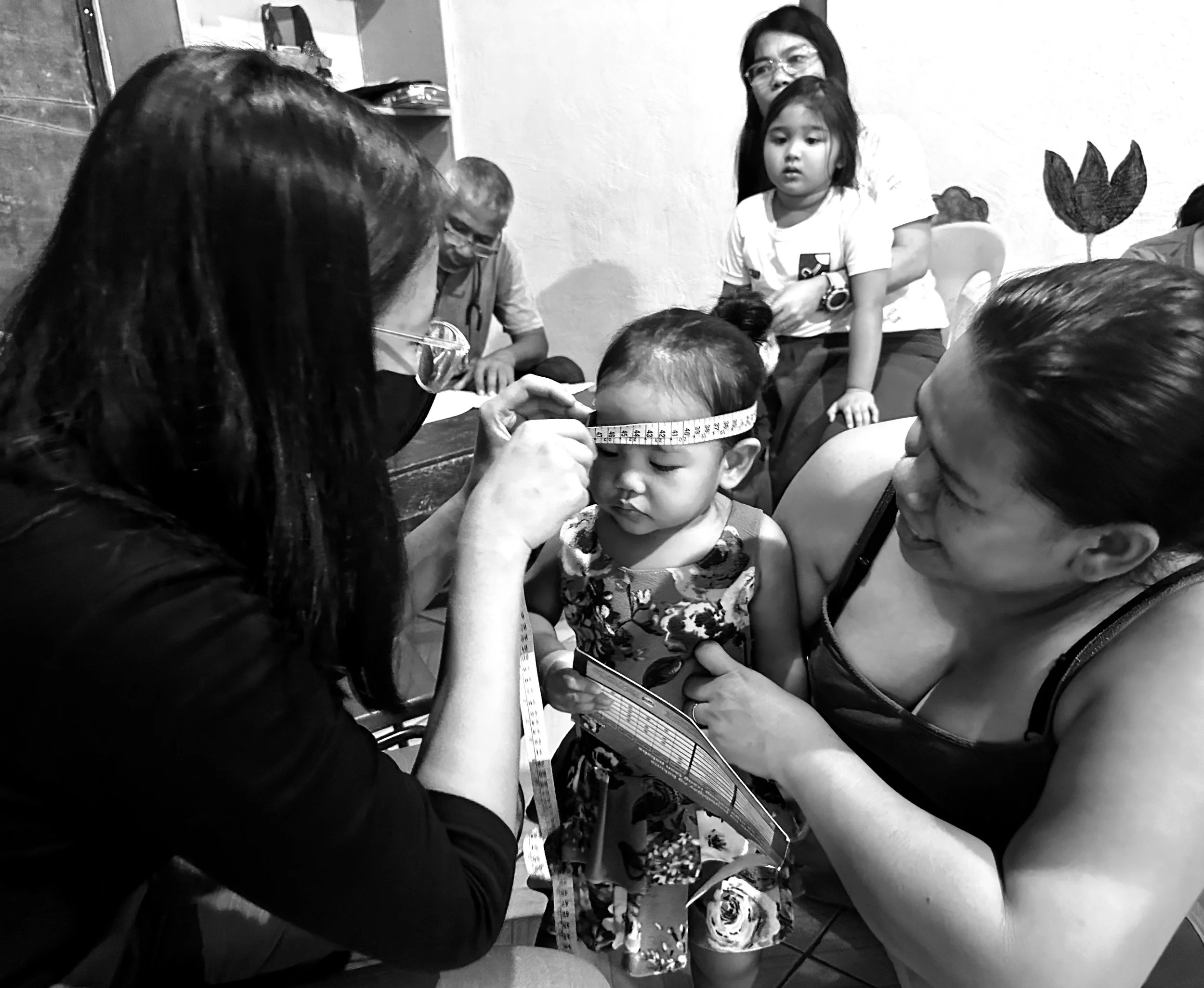 A woman measuring a child's head with a measuring tape while other children and adults observe, in a room with a painted flowering plant on the wall.©odoVita