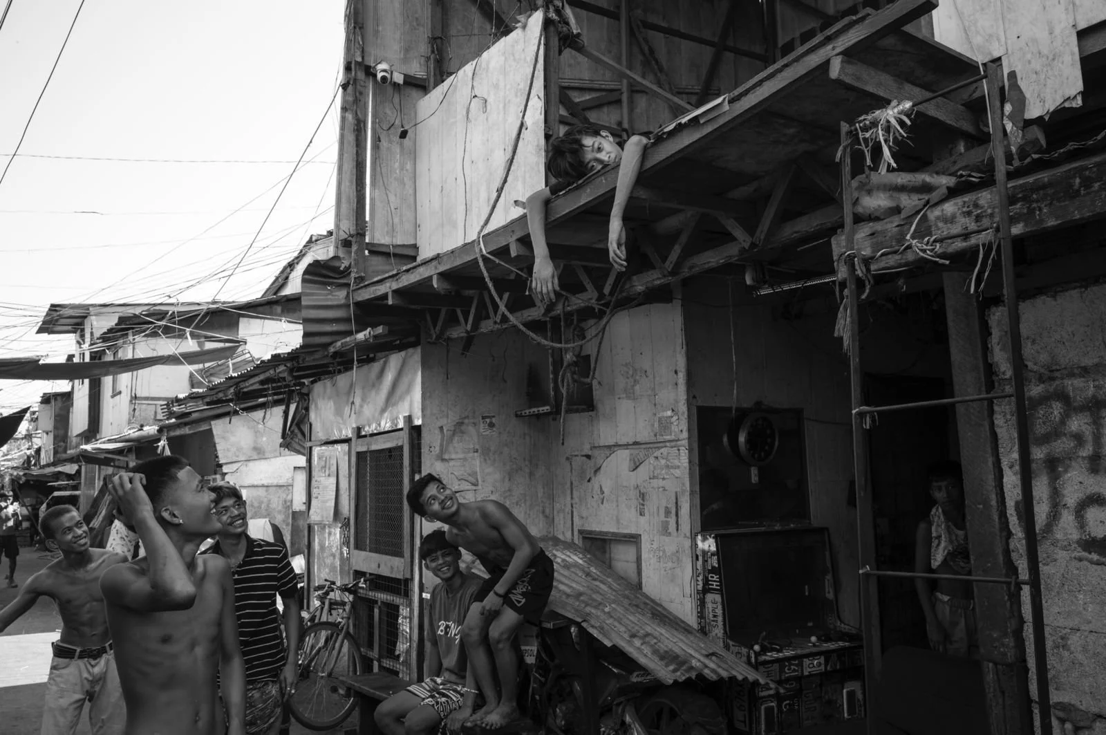 A group of children playing and laughing on a street in a neighborhood with makeshift houses. One boy is hanging from the balcony of a wooden house, while others look up and smile or laugh at him in Tondo, Manila ©R. Hartel for odoVita.