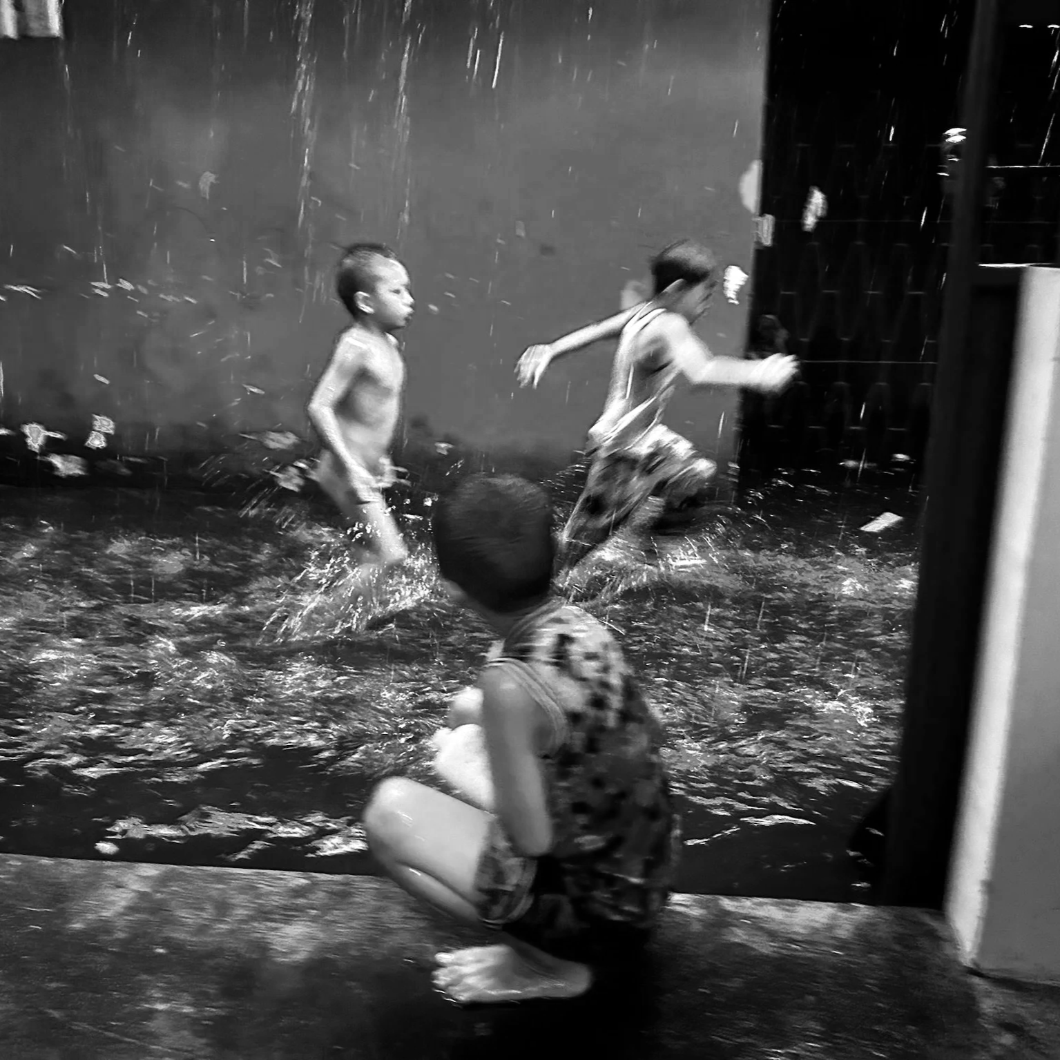 Two children are playing in the rain, one is running and the other is squatting with a container, surrounded by water splashing.©odoVita