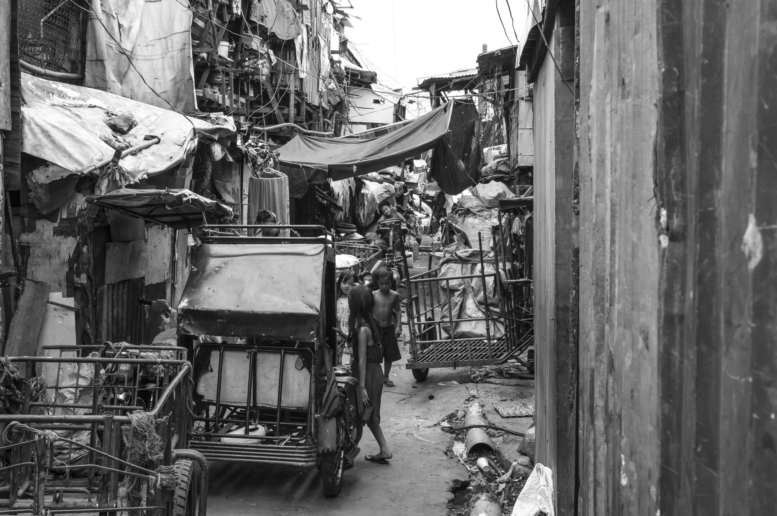 A narrow alley in a crowded, impoverished neighborhood with makeshift homes, children playing, and various items like carts and tarps scattered around in Tondo, Manila ©R. Hartel for odoVita.