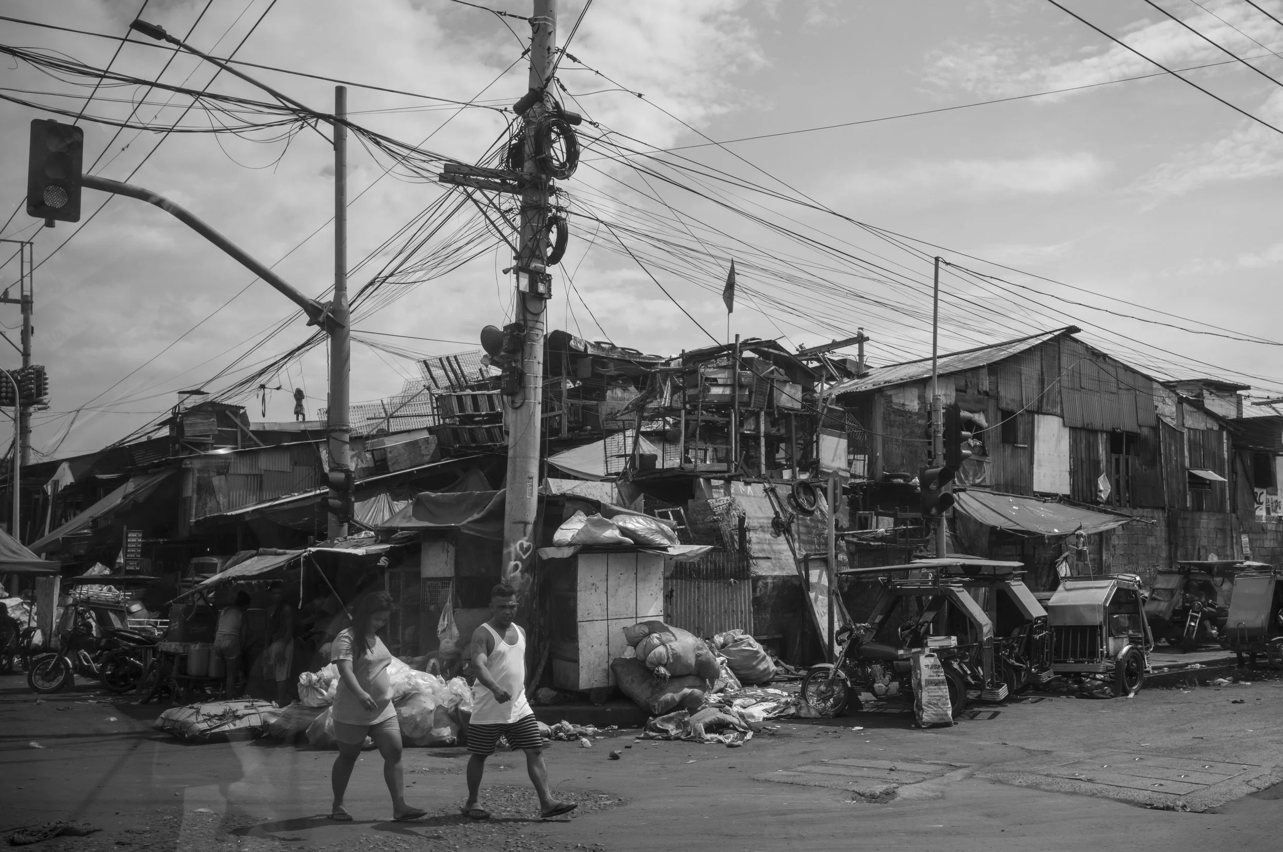 Black and white photo of a busy street scene with people walking past a makeshift marketplace. In the background, there are dilapidated buildings and a utility pole with numerous tangled electrical wires in Tondo, Manila ©R. Hartel for odoVita.
