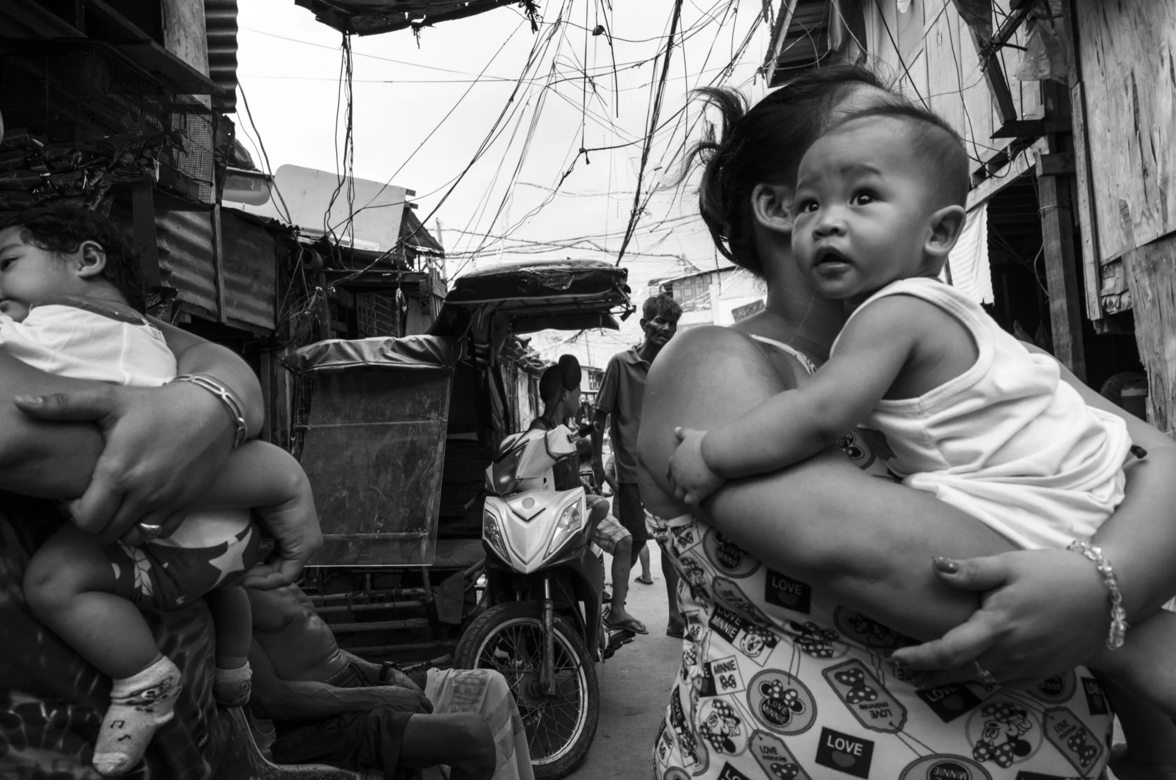 A woman holding a young child in a crowded street with makeshift houses and electrical wires overhead.©R. Hartel for odoVita